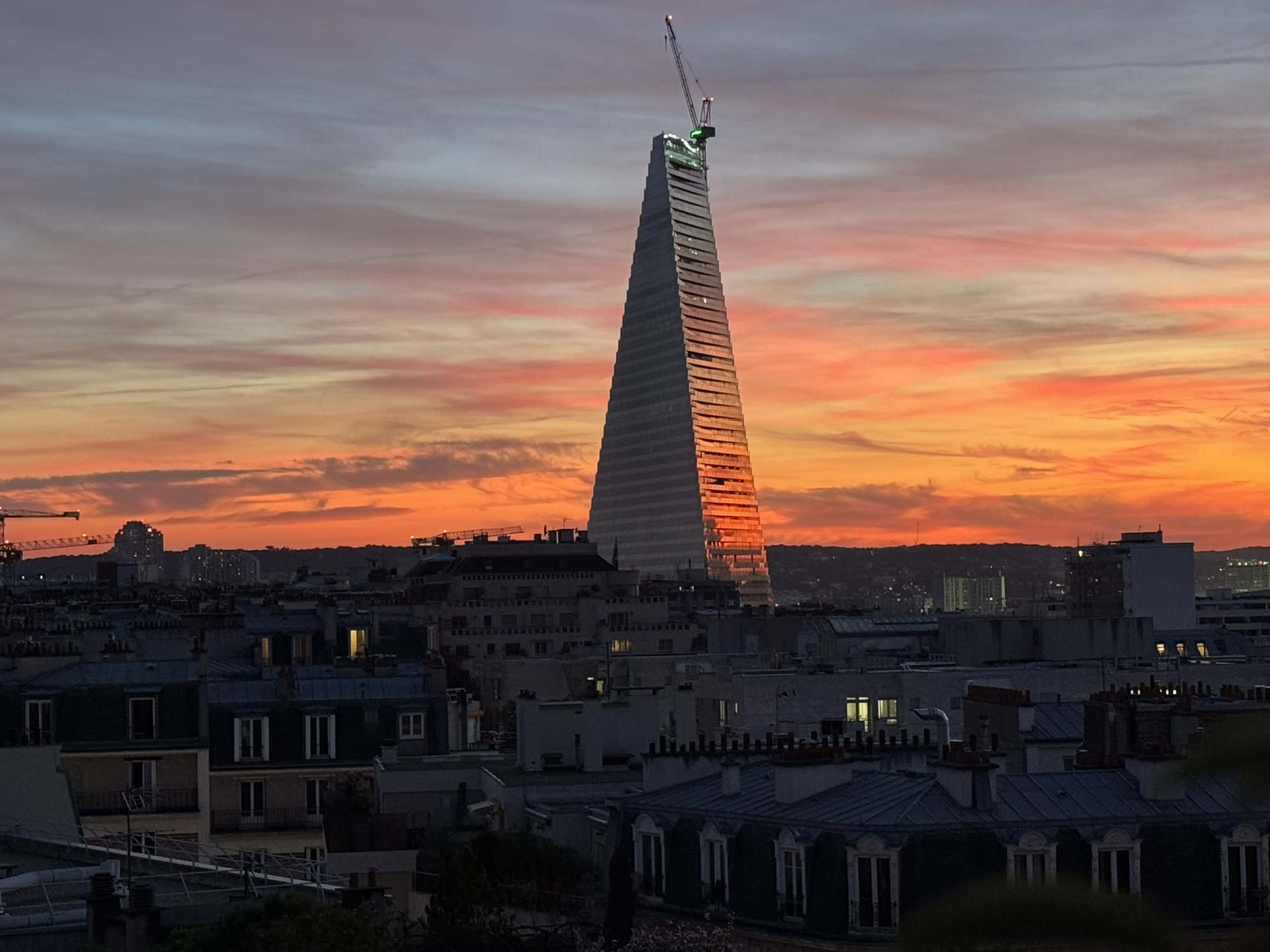 The image shows a tall, modern skyscraper with a triangular shape, illuminated by a vibrant sunset in the background.