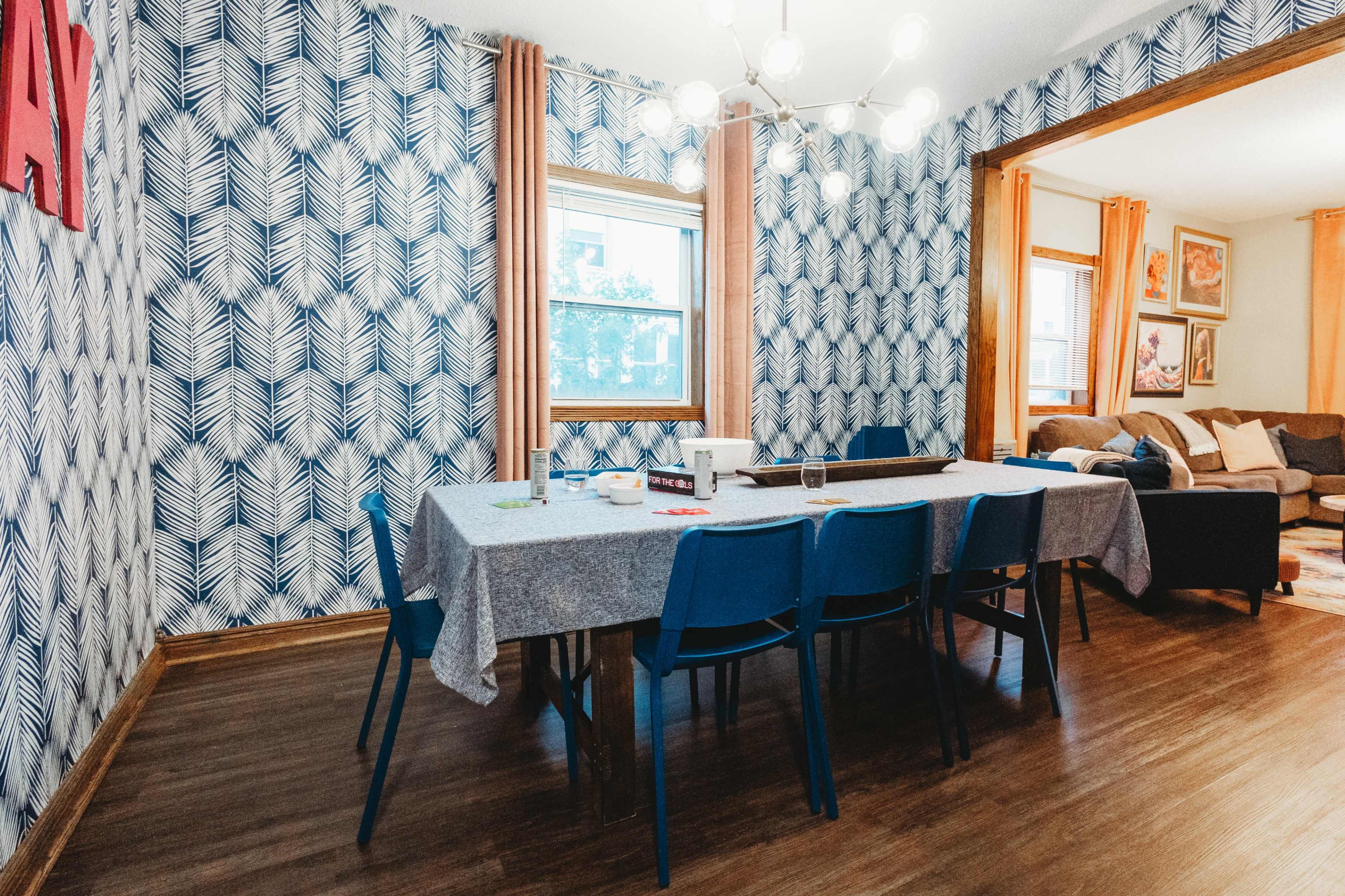 A dining area features a large table surrounded by blue chairs, with patterned wallpaper and a living room visible in the background.
