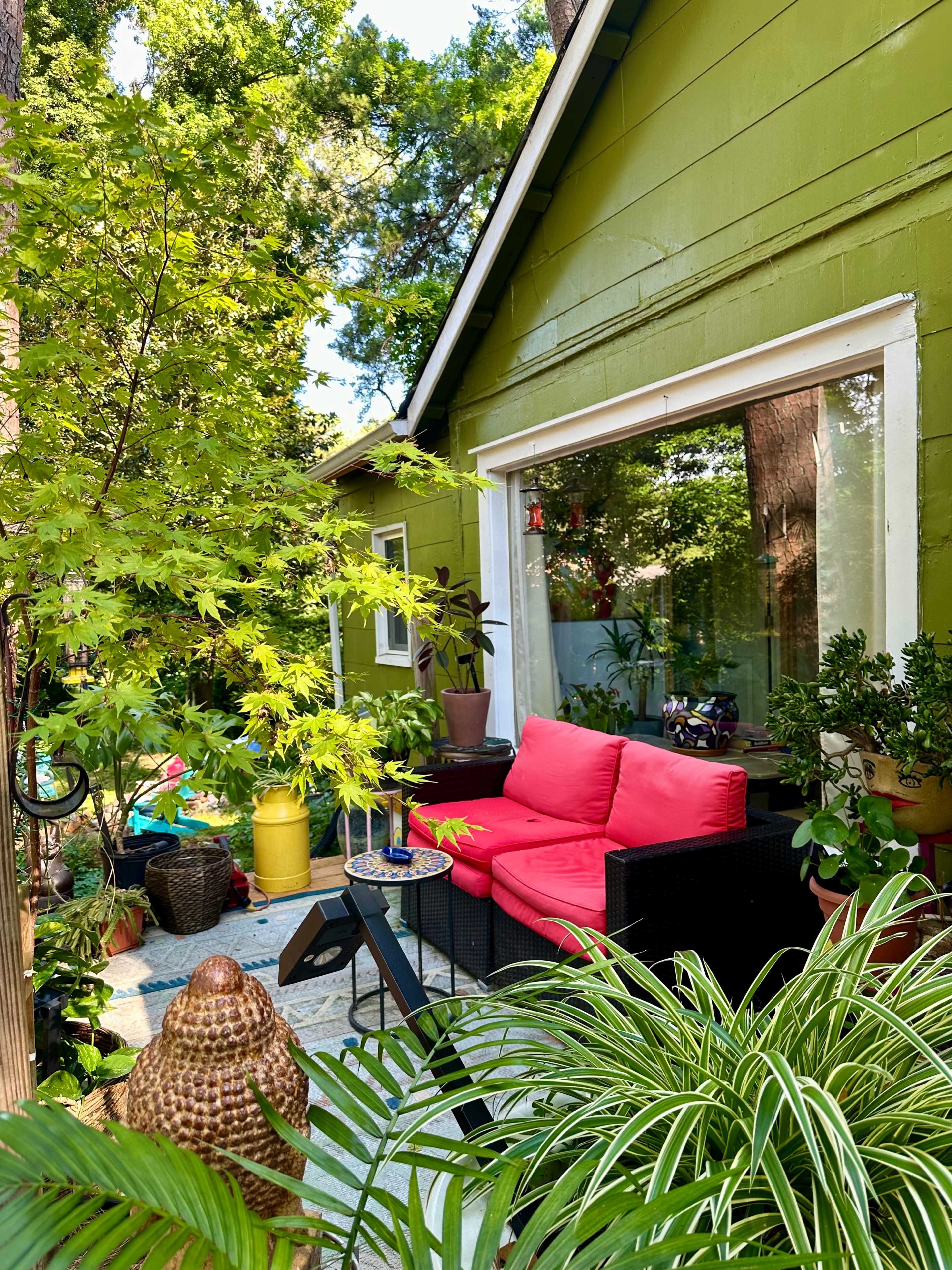 A green house is flanked by lush greenery and features a small patio with a pink couch and potted plants.