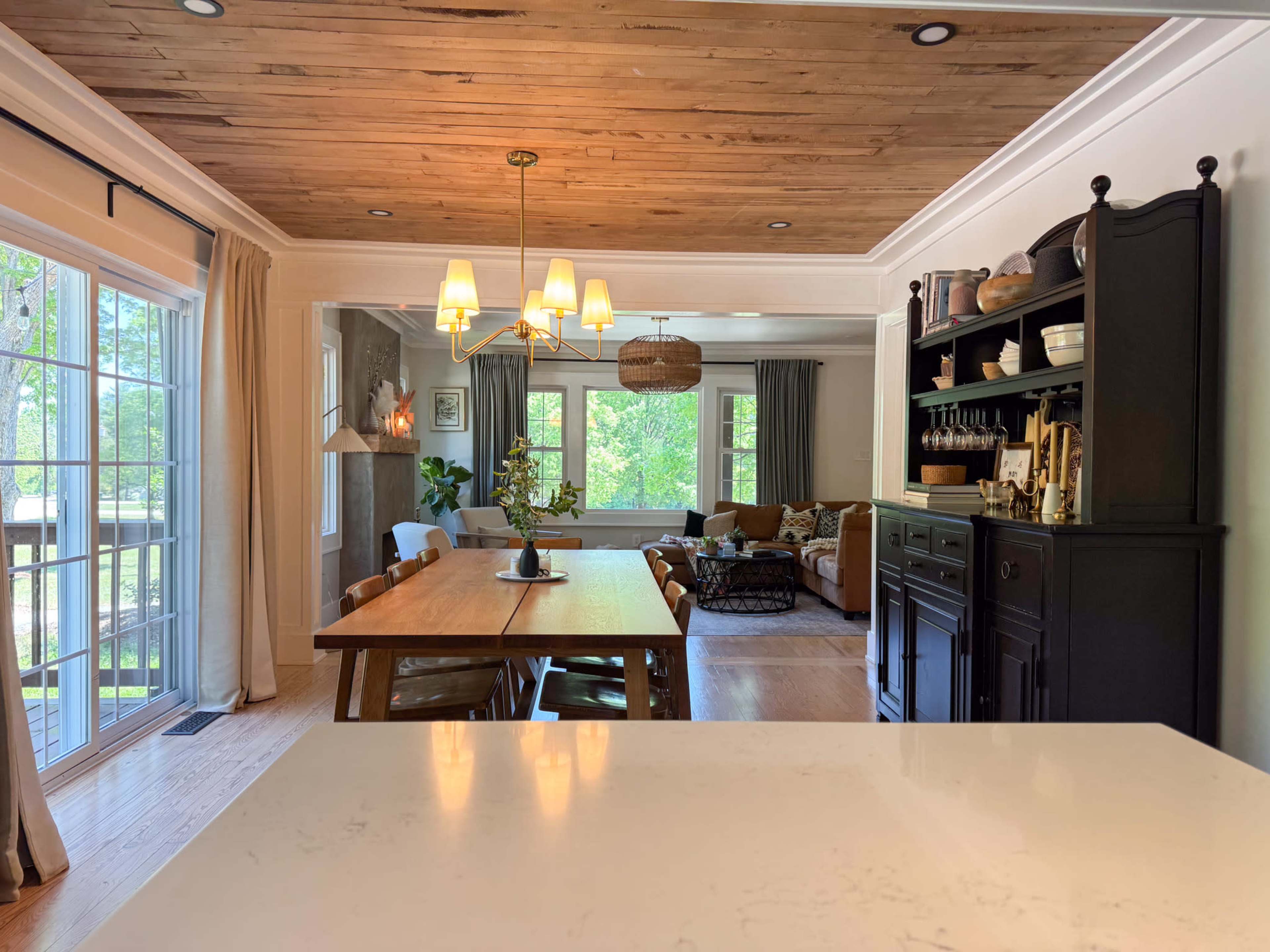 The image shows a dining area with a wooden table, a black cabinet, and a living room in the background featuring a large window, sofa, and decorative elements.