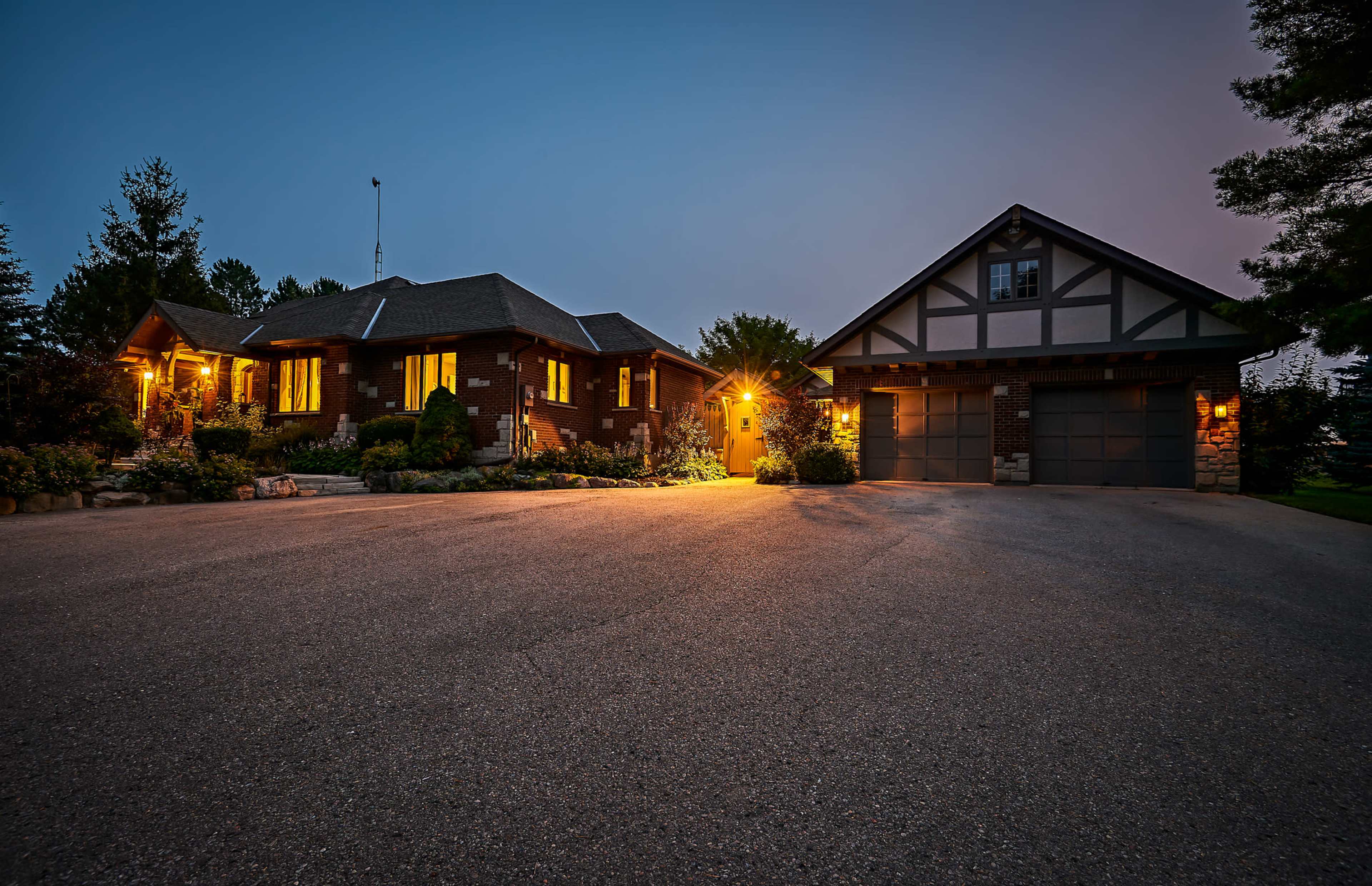 The image shows a well-lit brick house with large windows and a separate garage, surrounded by trees at dusk.