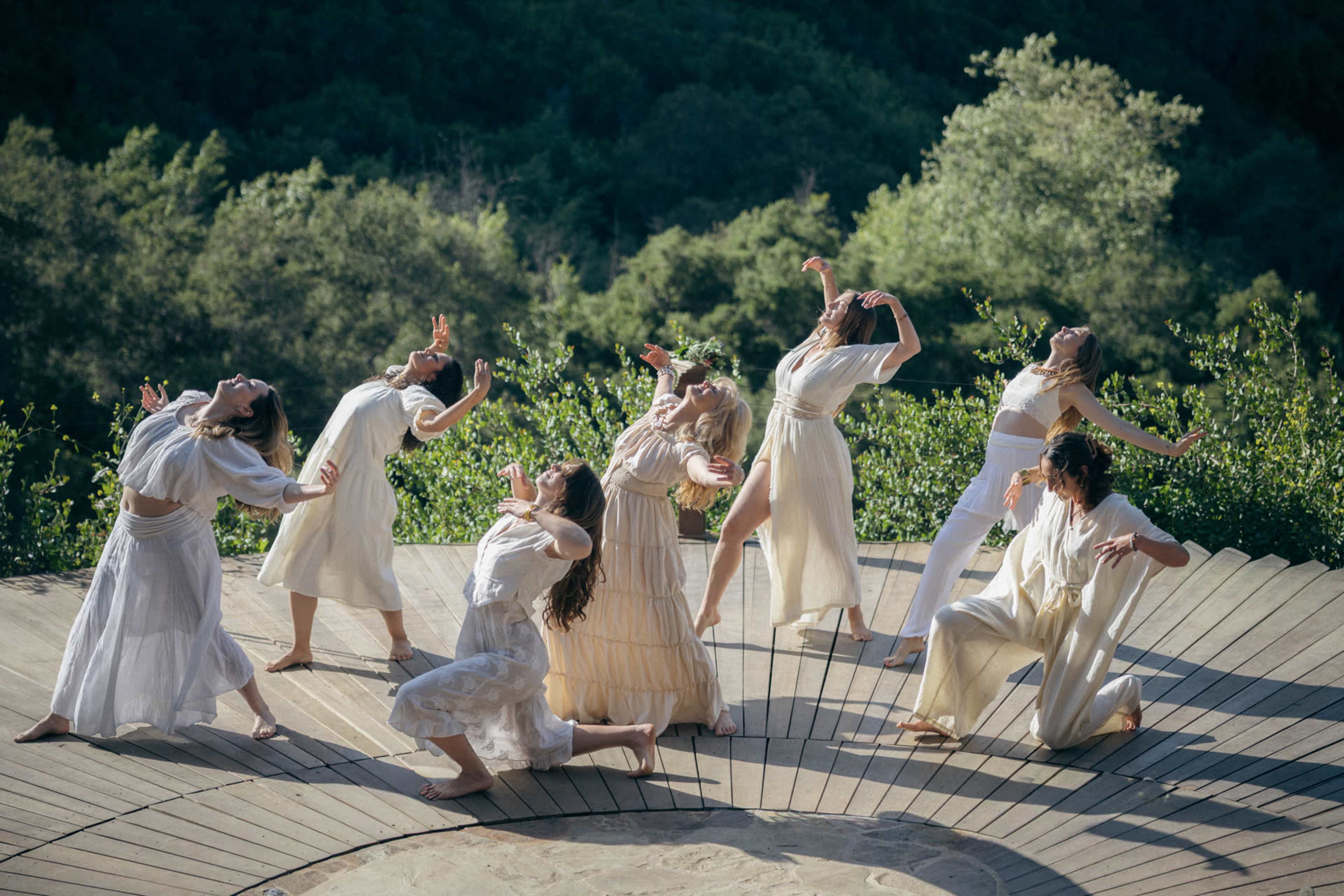 A group of eight dancers in white clothing is performing on a circular wooden stage surrounded by greenery.