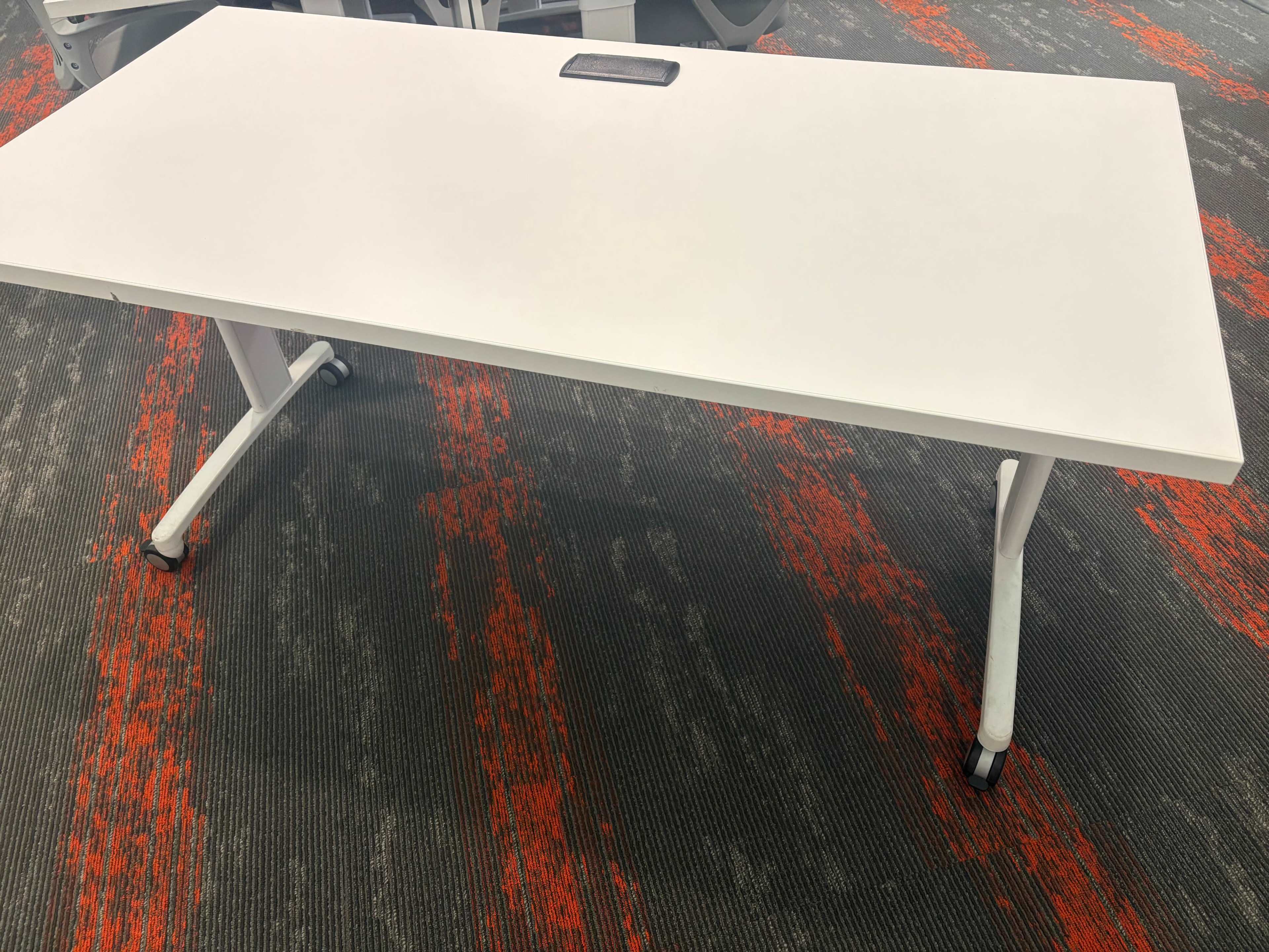 A white desk with a surface mount and wheeled legs is placed on a patterned gray and orange carpet.