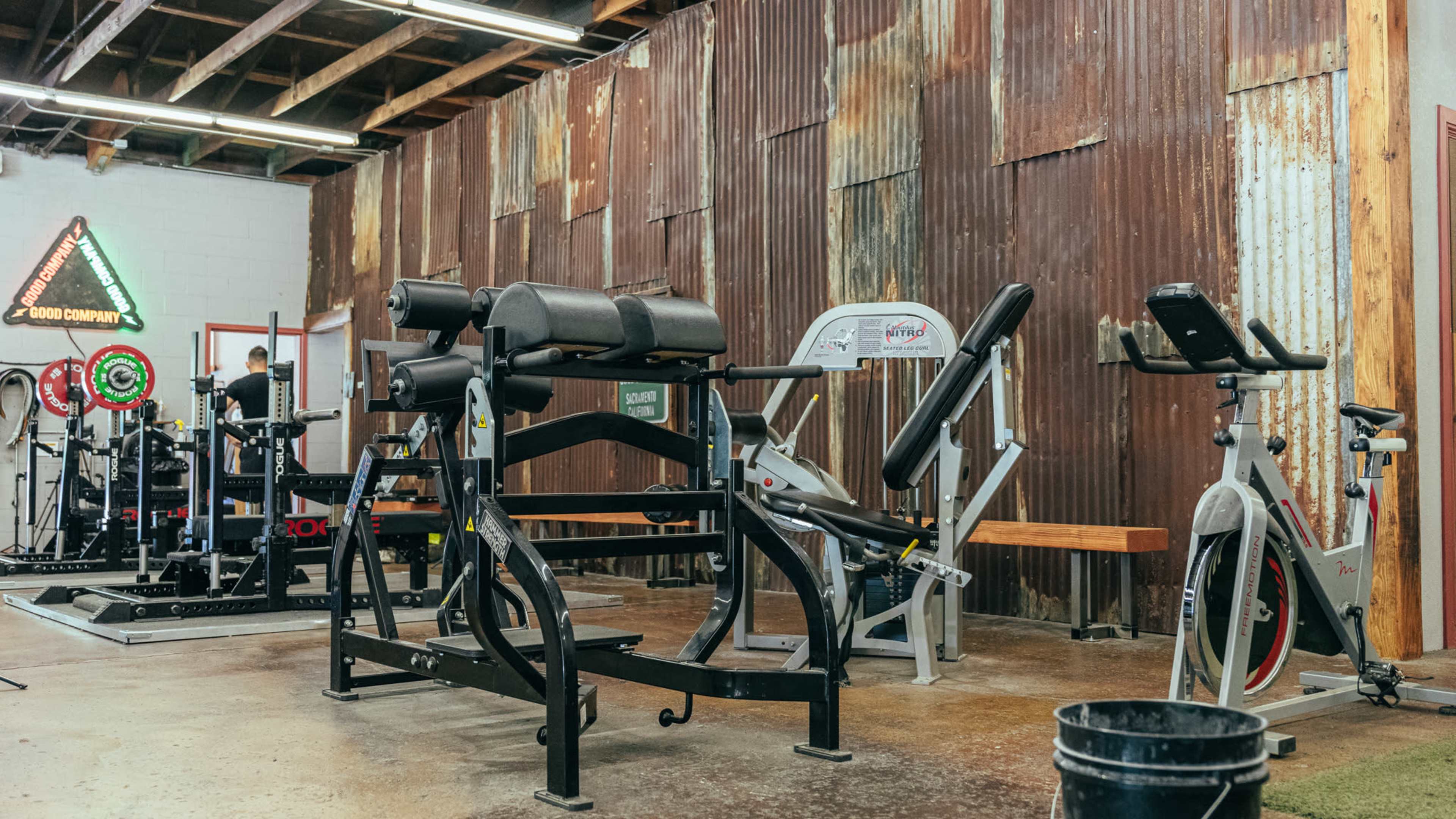 A gym interior featuring various workout equipment, including benches, exercise machines, and stationary bicycles against a rustic metal wall.
