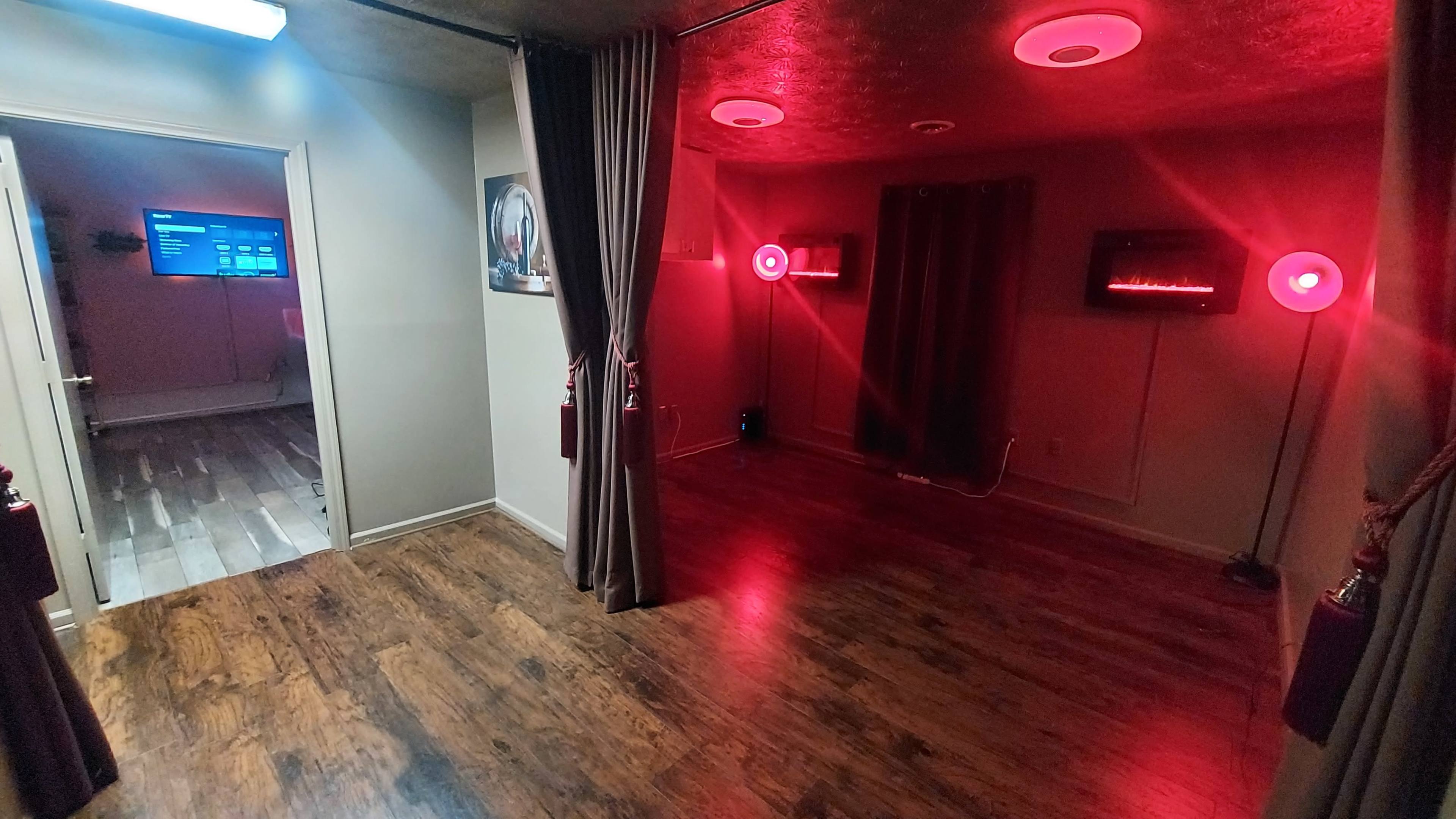 The image shows a dimly lit room with wooden flooring, red lighting from circular fixtures, and two screens mounted on the walls, flanked by curtains.