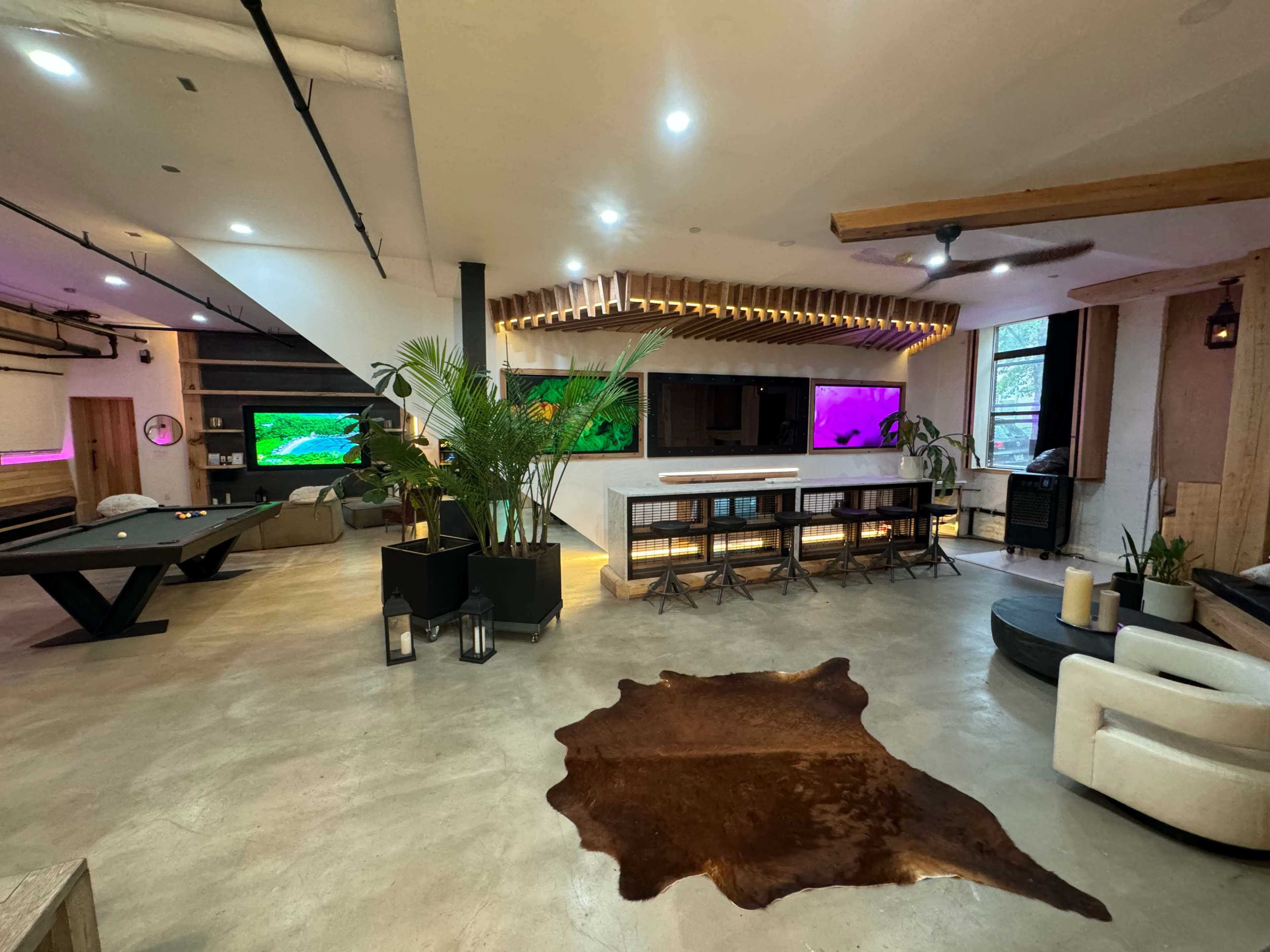 The image shows a spacious, modern living area with a pool table, illuminated wall decorations, and large windows, featuring a combination of natural elements and contemporary design.