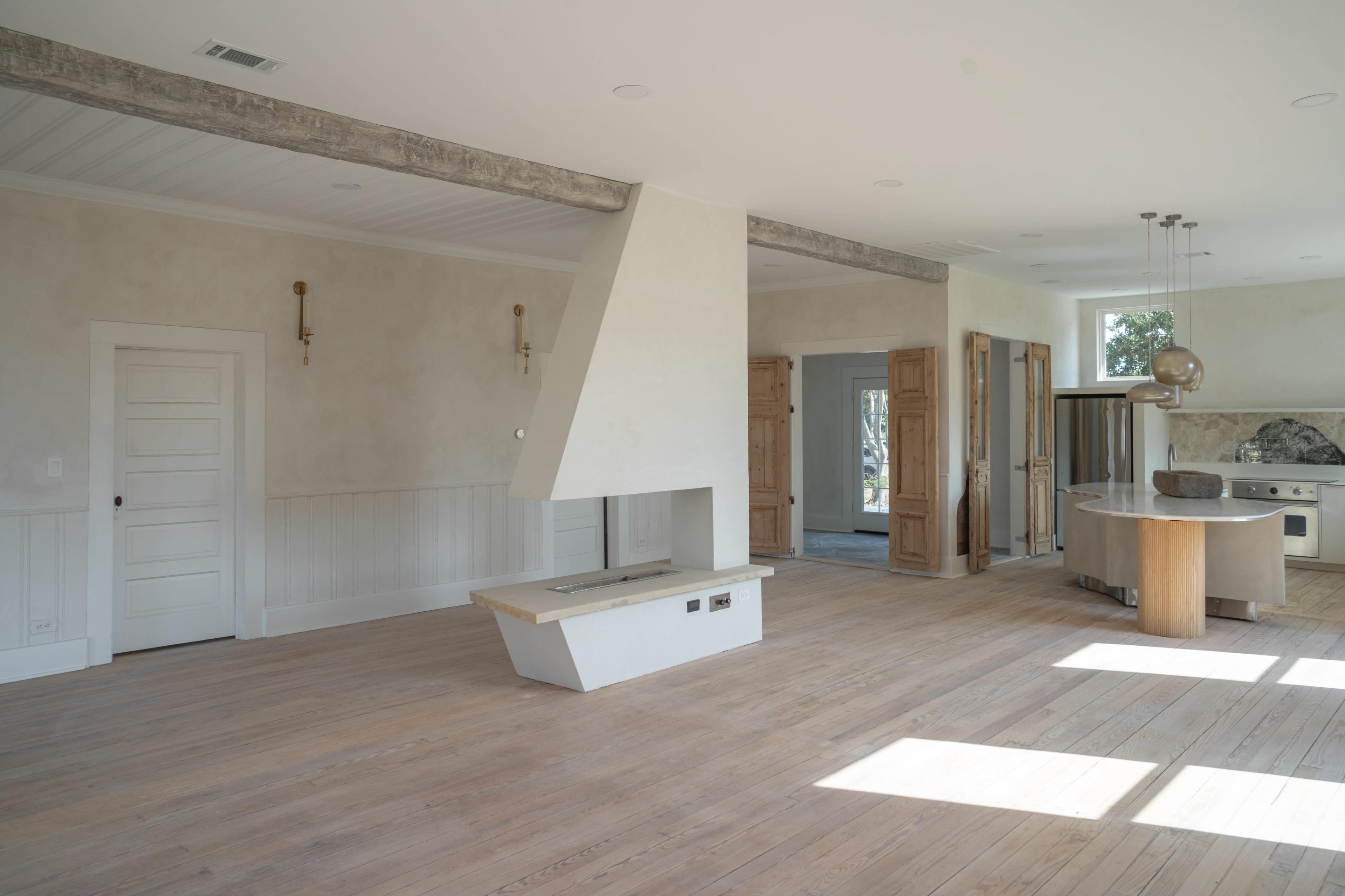 A spacious, modern interior of a room with wooden floors, a central fireplace, and an open layout connecting a kitchen and additional rooms.