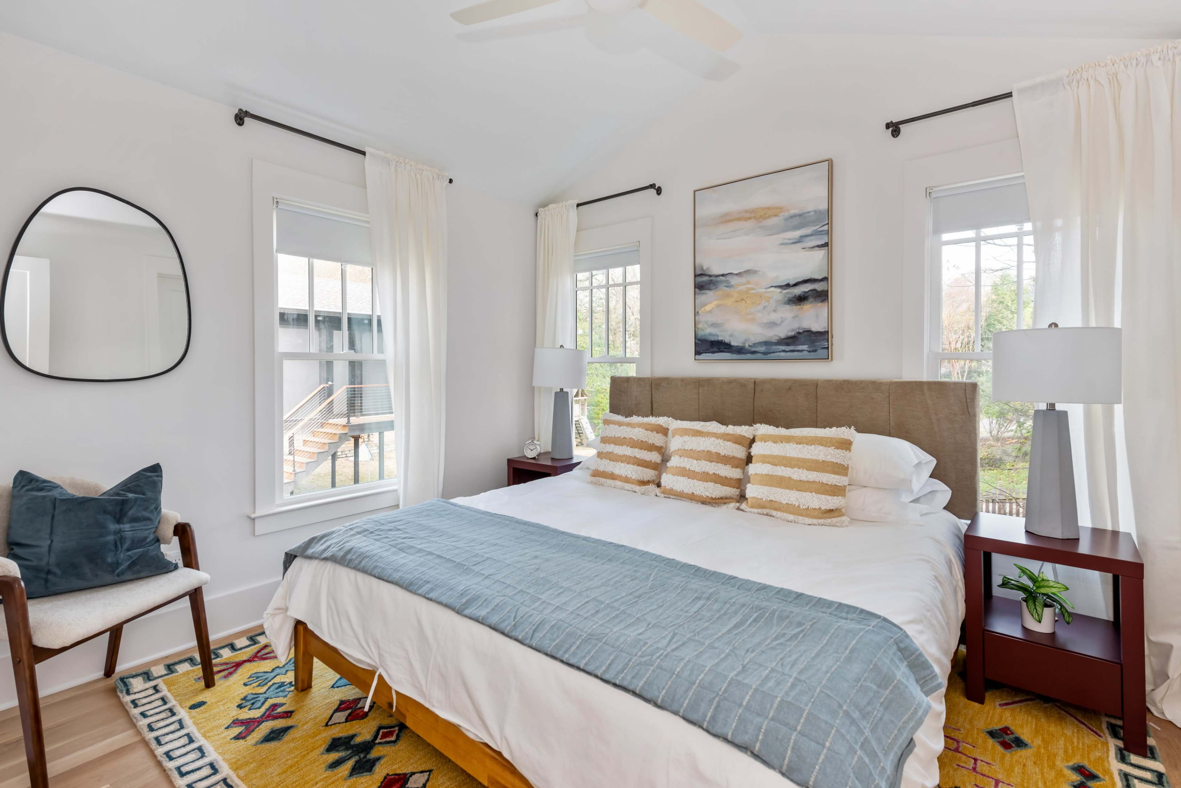 A bright bedroom features a large bed with decorative pillows, a mirror on the wall, and windows overlooking an outdoor area, with simple furnishings and a colorful rug.
