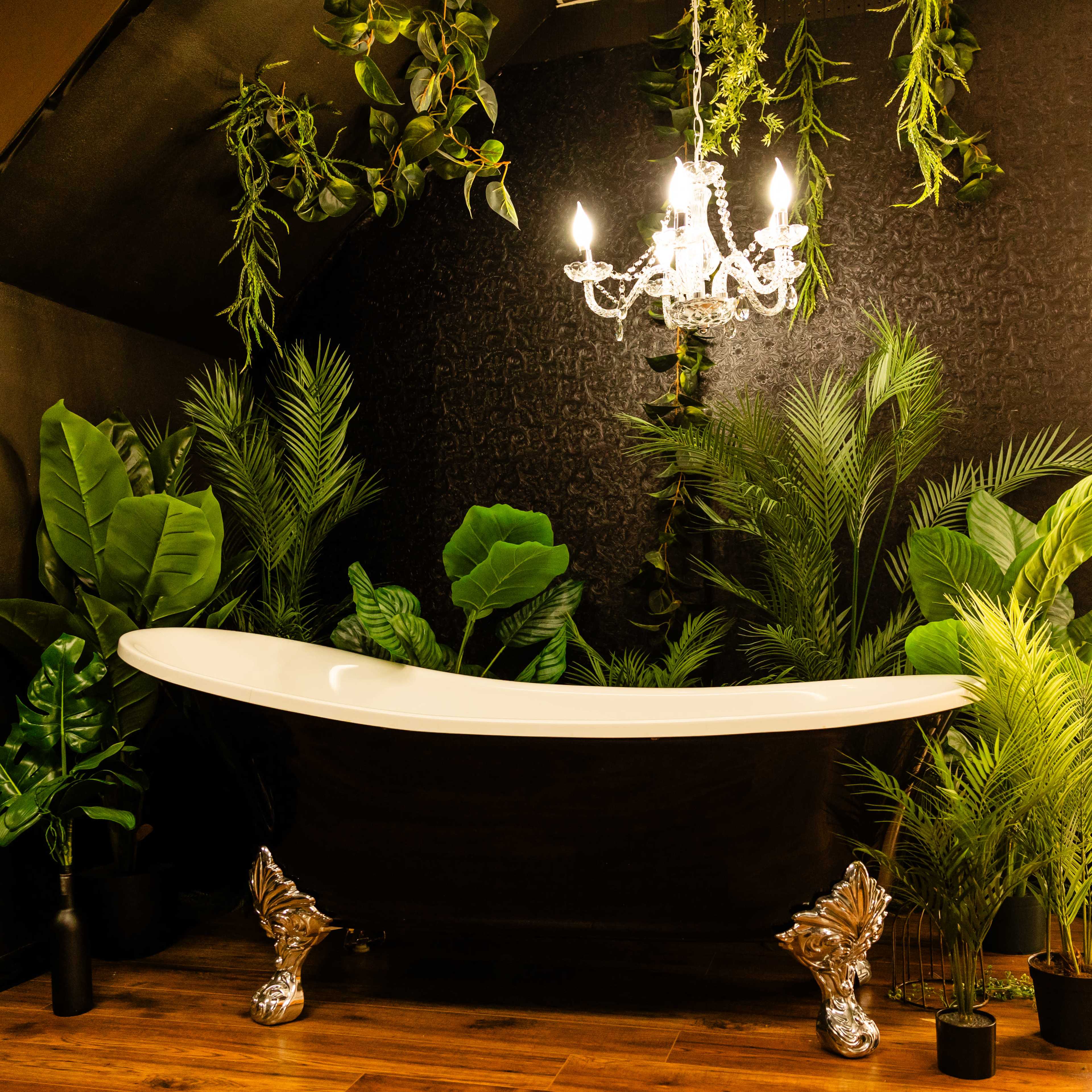 A black freestanding bathtub with silver legs is surrounded by lush green plants and hangs a chandelier from the ceiling.