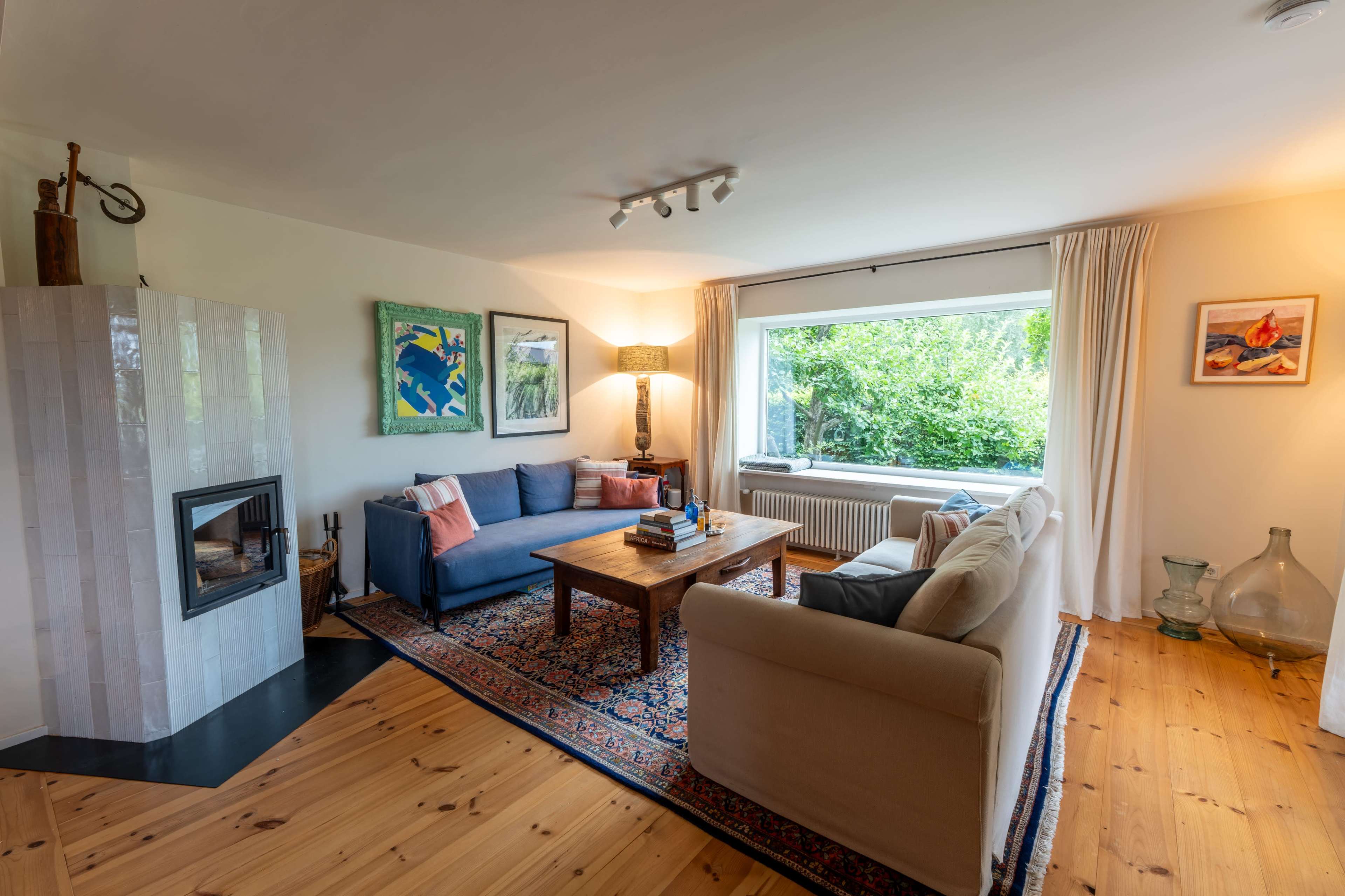 A cozy living room featuring a blue sofa, a beige couch, a wooden coffee table, and a fireplace, with large windows providing a view of greenery outside.