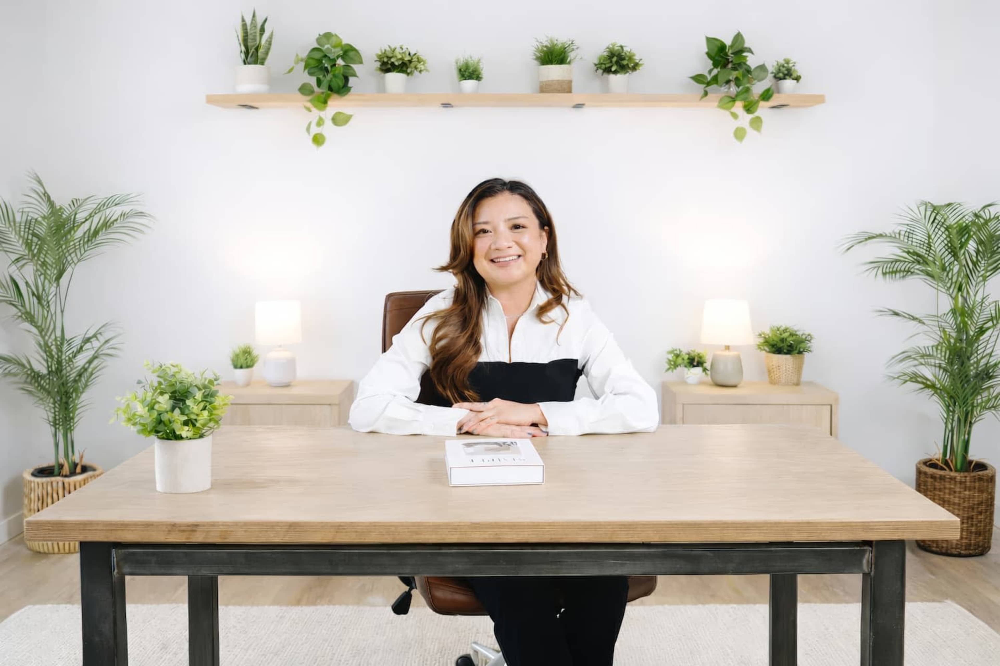 A woman sits at a wooden desk in a modern office space decorated with plants and soft lighting.