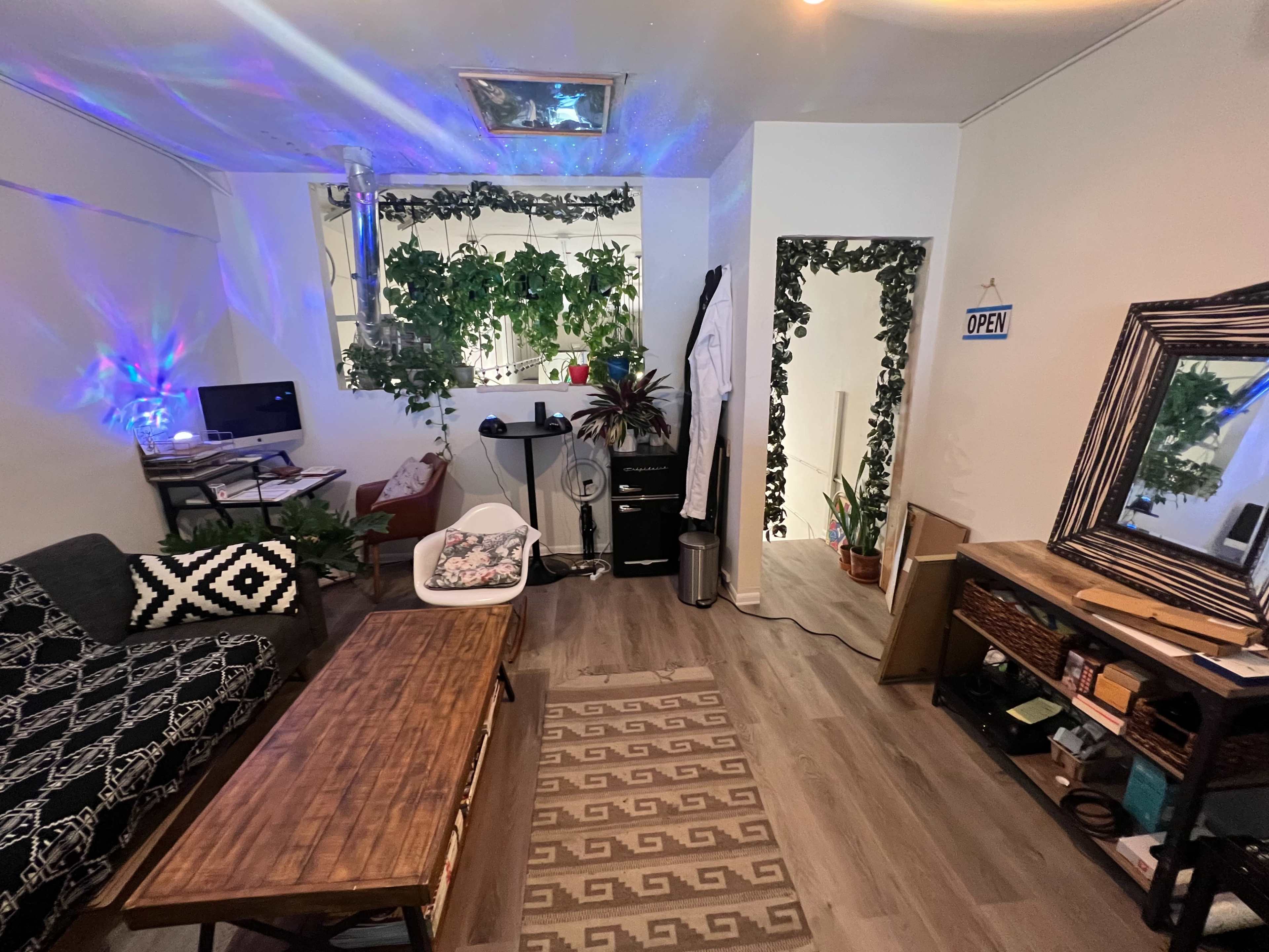 A living space with a couch, a wooden table, a computer setup, plants on the walls, and an open doorway leading to another room.