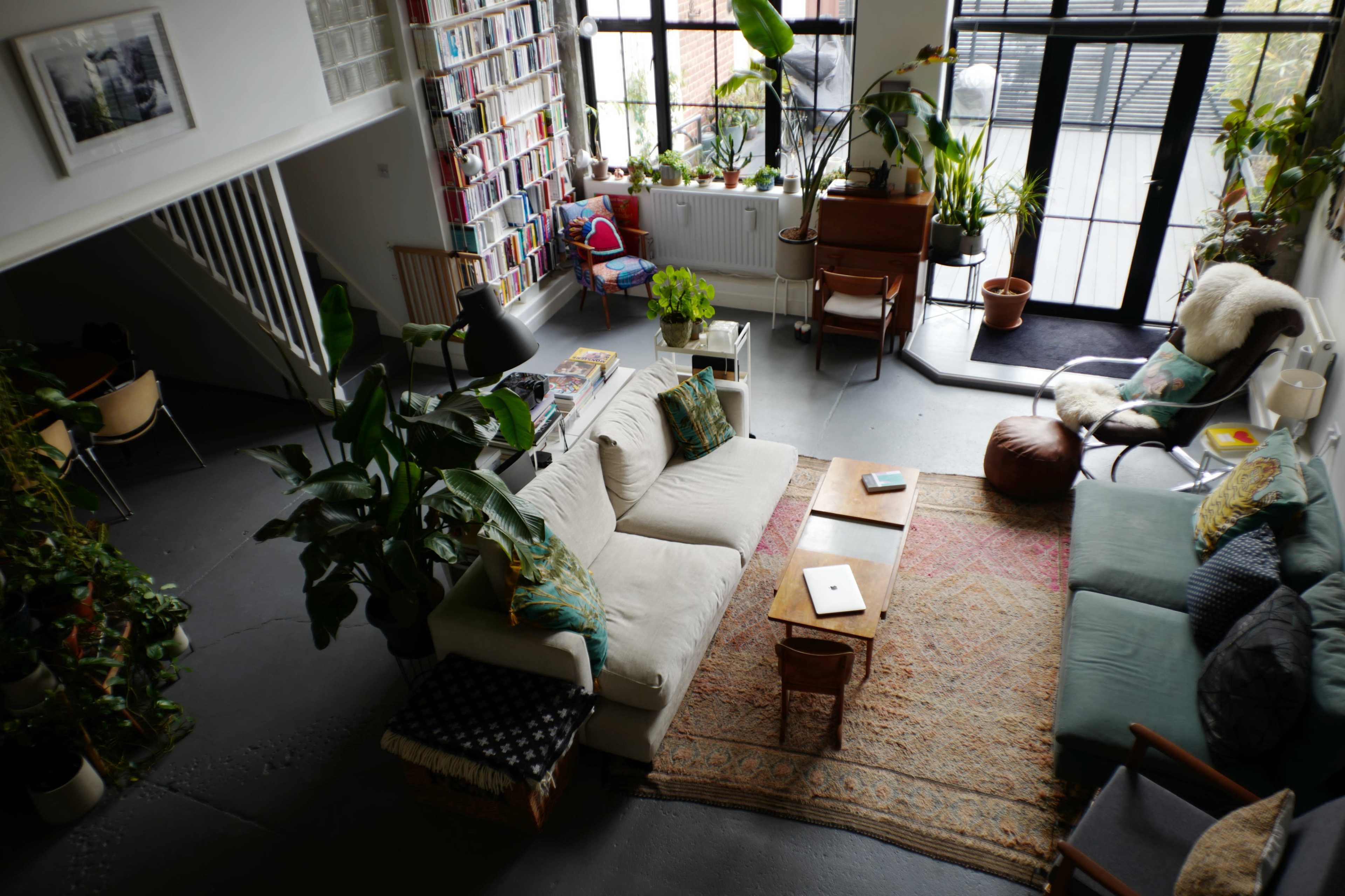 The image shows a spacious living room filled with plants, a large sofa, a coffee table, and a bookshelf, with large windows allowing natural light to enter.