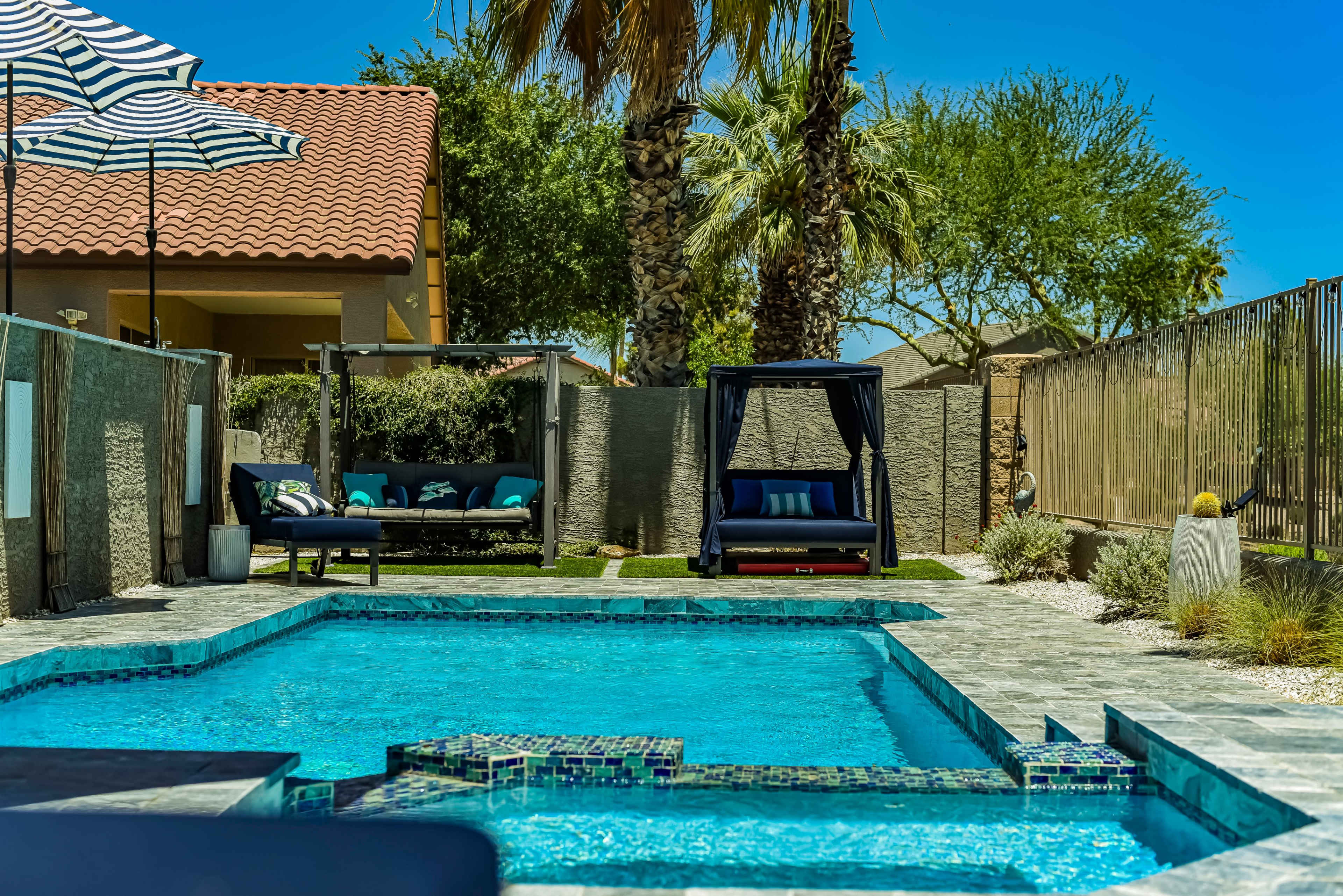 A residential backyard featuring a swimming pool, lounge chairs, a swinging daybed, and palm trees under a clear blue sky.