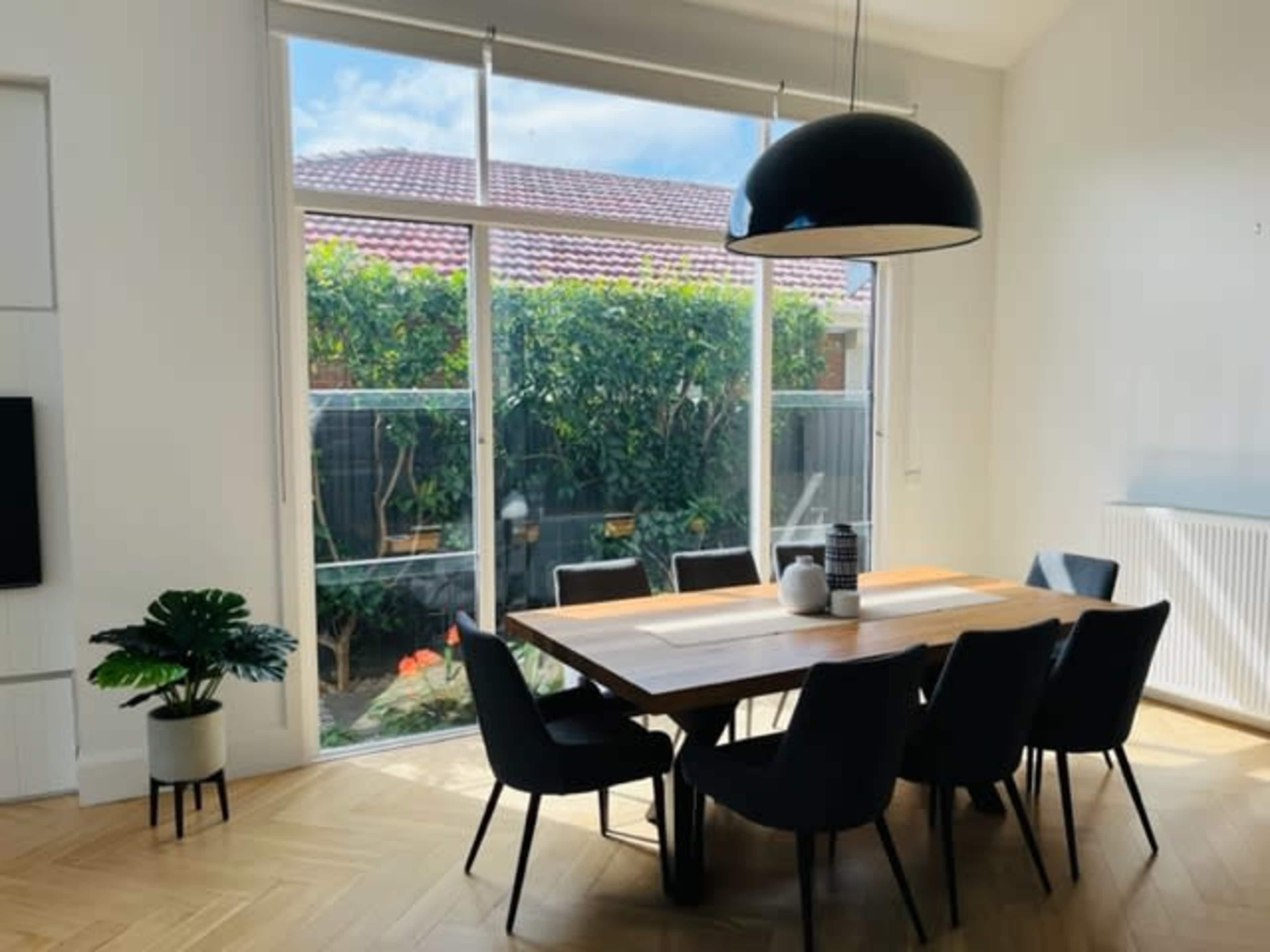 A dining area features a large wooden table surrounded by black chairs, with a view of a garden visible through large windows.