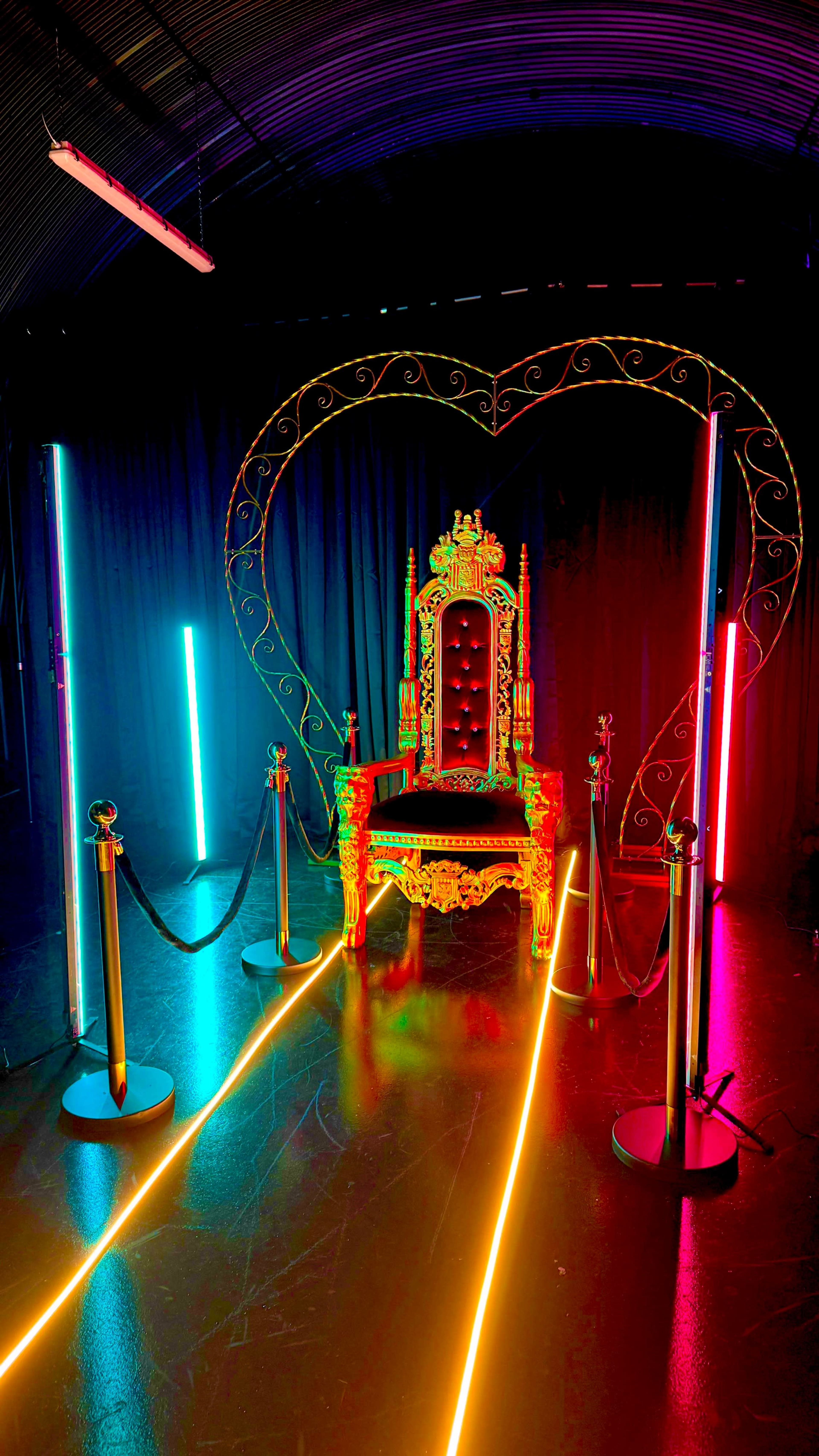 The image shows an elaborate throne illuminated by colorful neon lights in a darkened room, surrounded by velvet ropes and light stands.