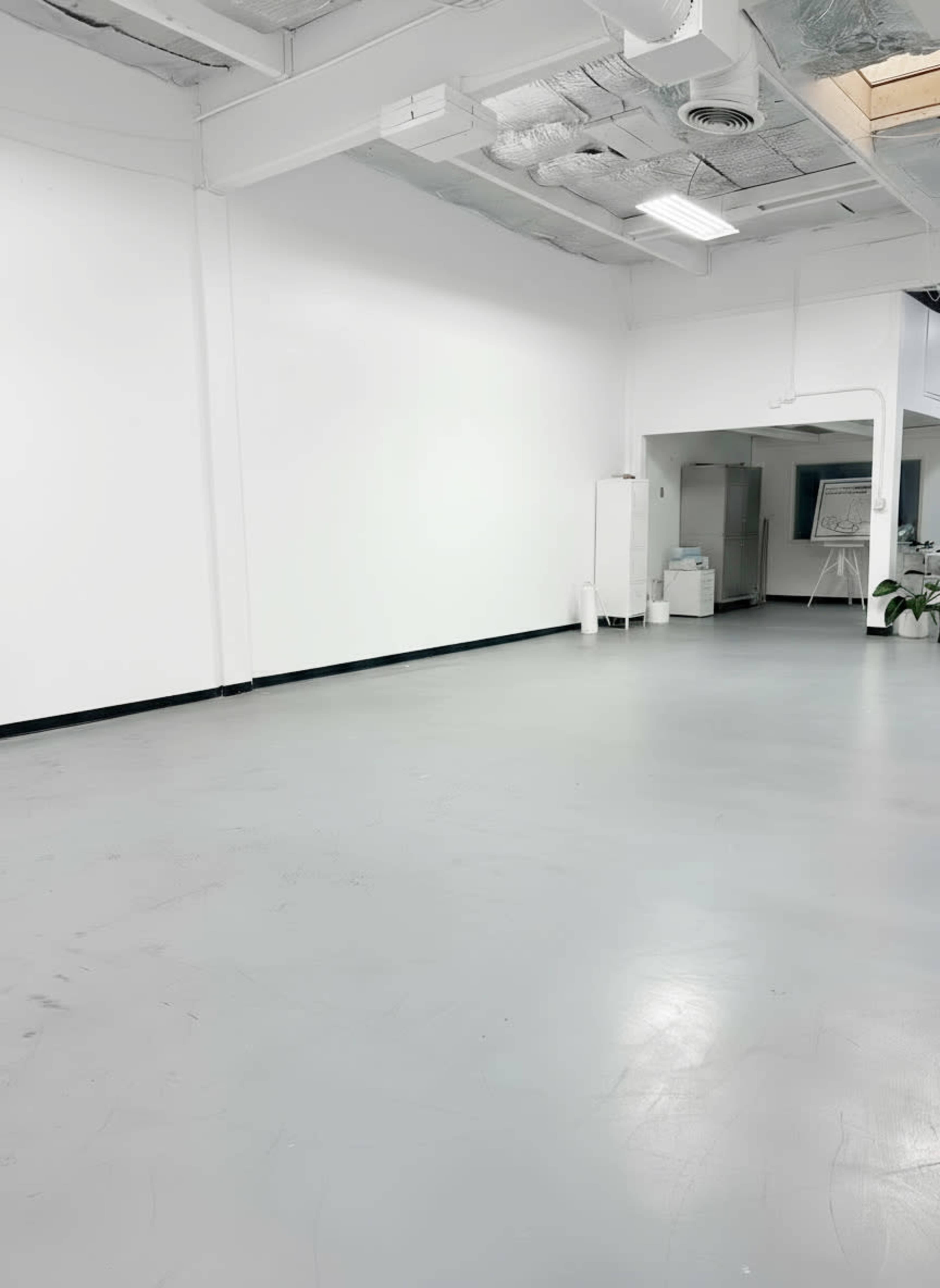 The image shows an empty, well-lit room with gray flooring and white walls, featuring a doorway leading to another space.