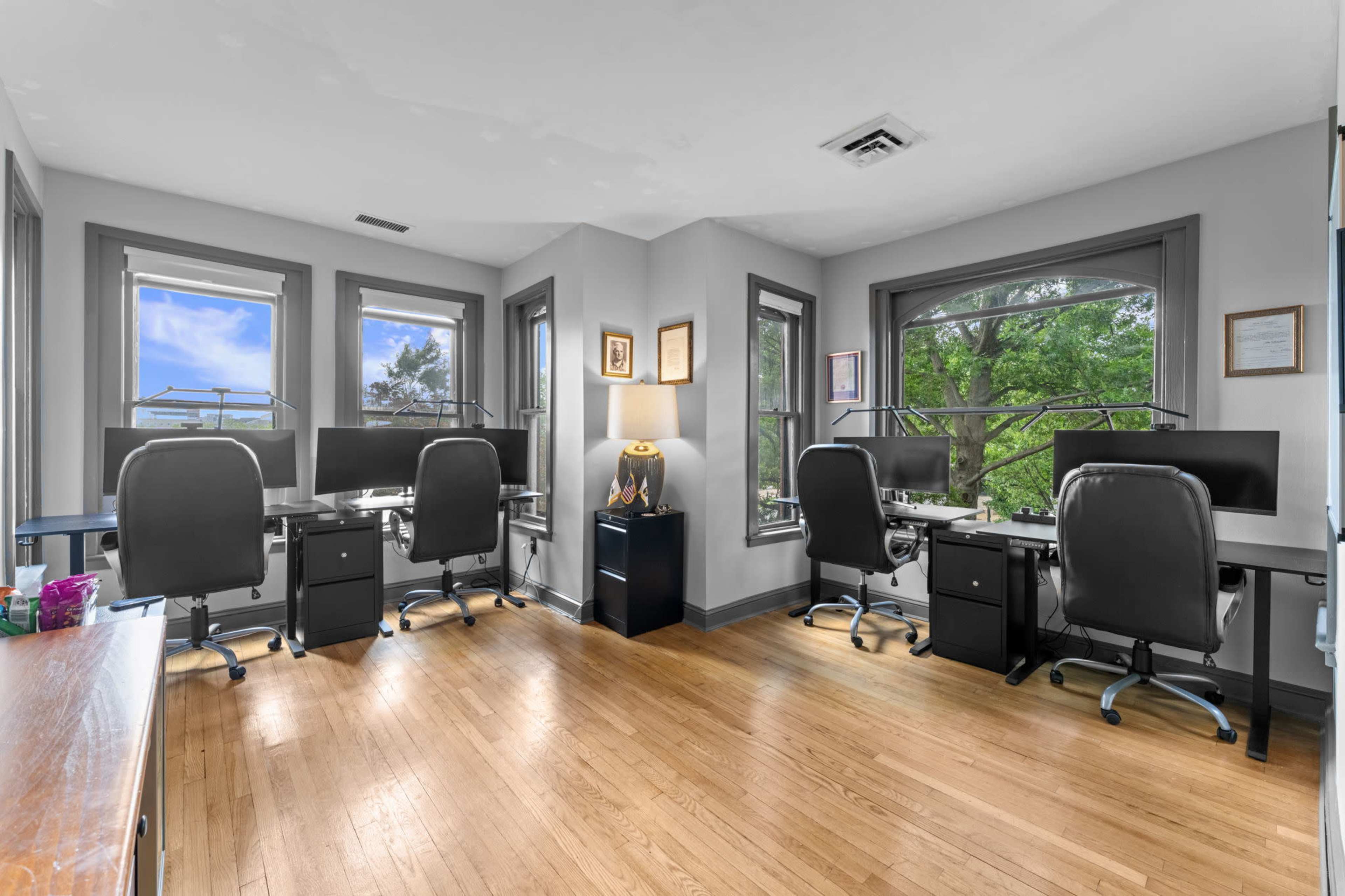 The image shows a bright office space with four desks, each equipped with computer setups, arranged around large windows that offer views of greenery.