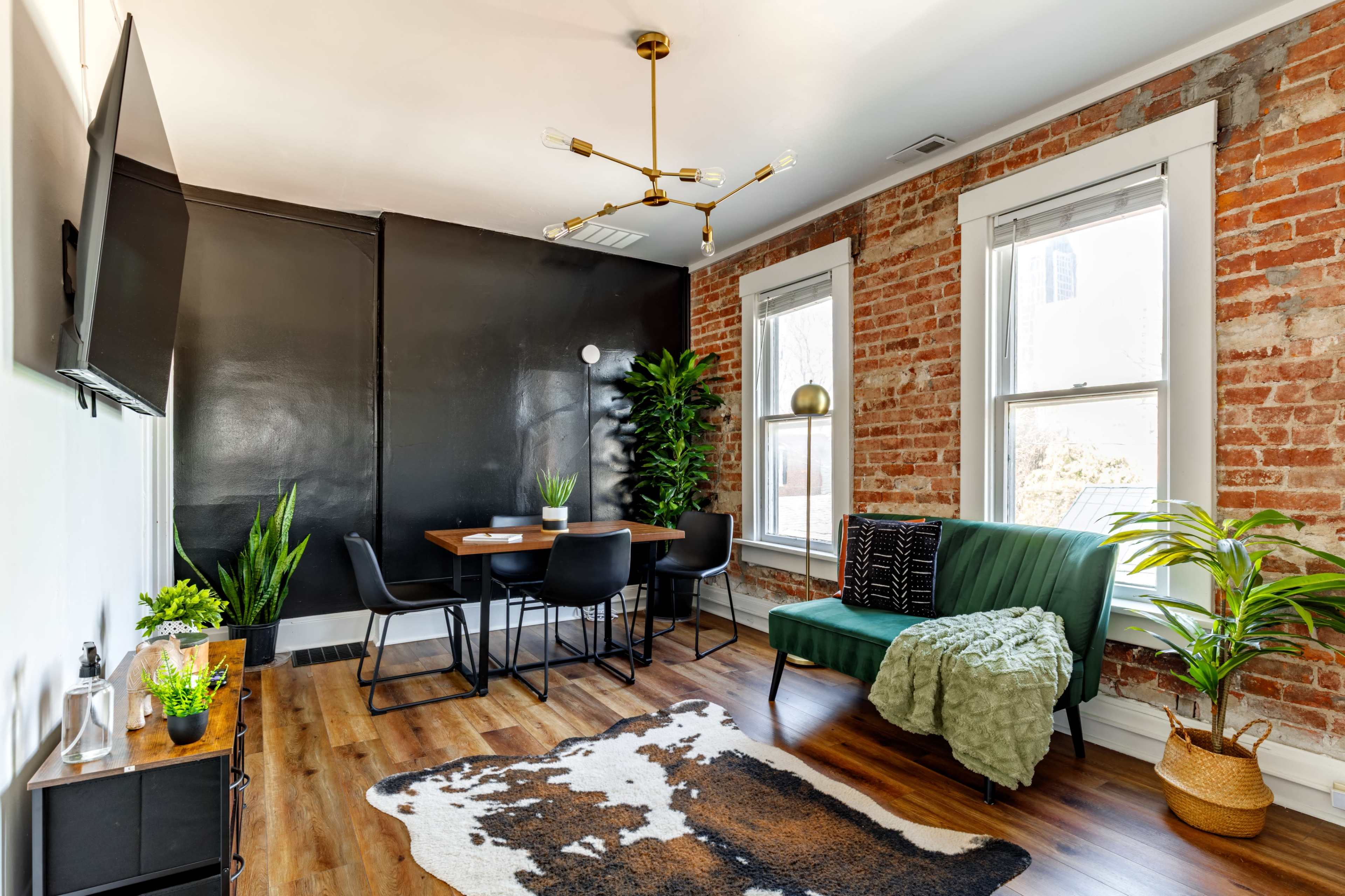 The image shows a modern living space featuring exposed brick walls, a green sofa, a dining table with chairs, and decorative plants.