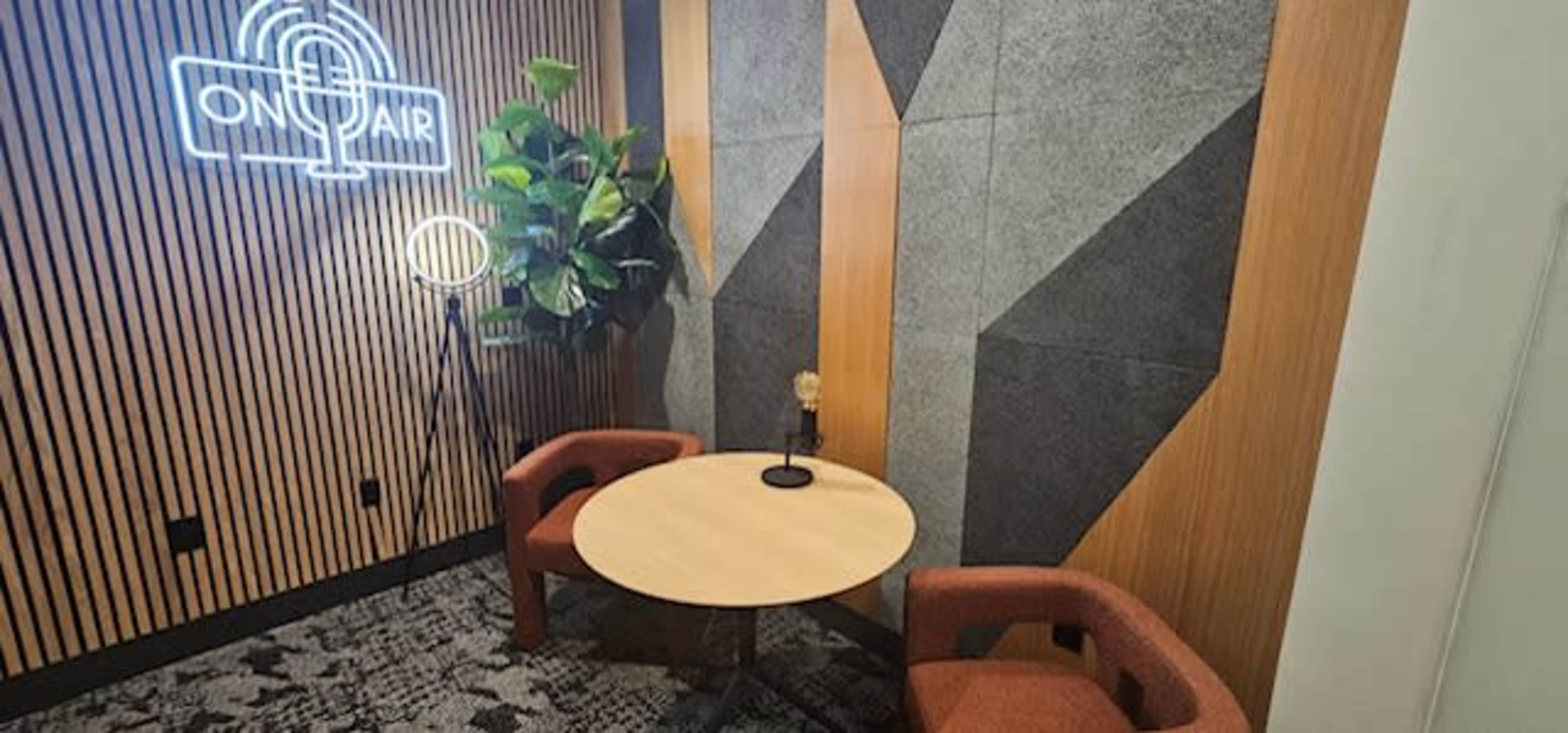The image shows a small, modern recording or meeting space with a round table, two upholstered chairs, a decorative plant, and a neon "ON AIR" sign on the wall.