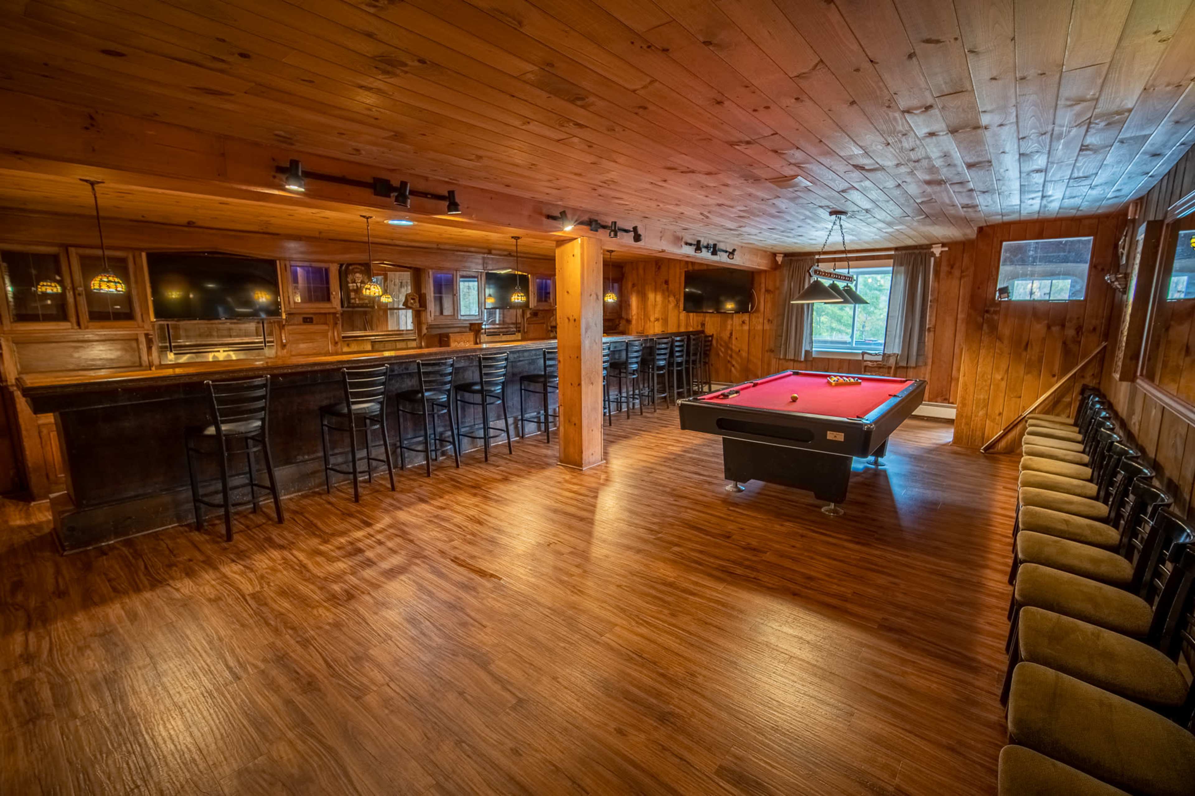 A wooden-paneled recreation room featuring a pool table, a bar area with stools, and several chairs lined against the wall.
