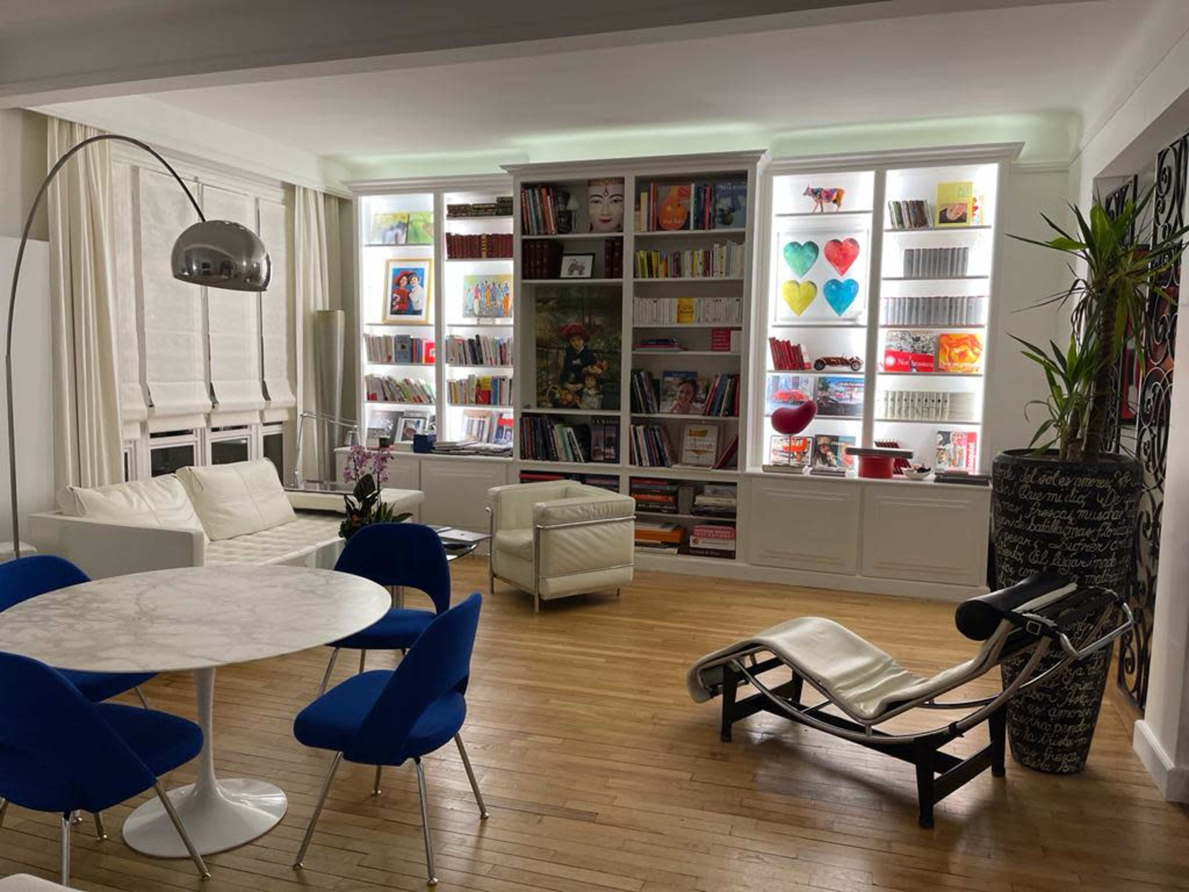 A modern living space features a circular marble dining table surrounded by blue chairs, with white sofas and a large bookshelf displaying colorful artwork and decor.