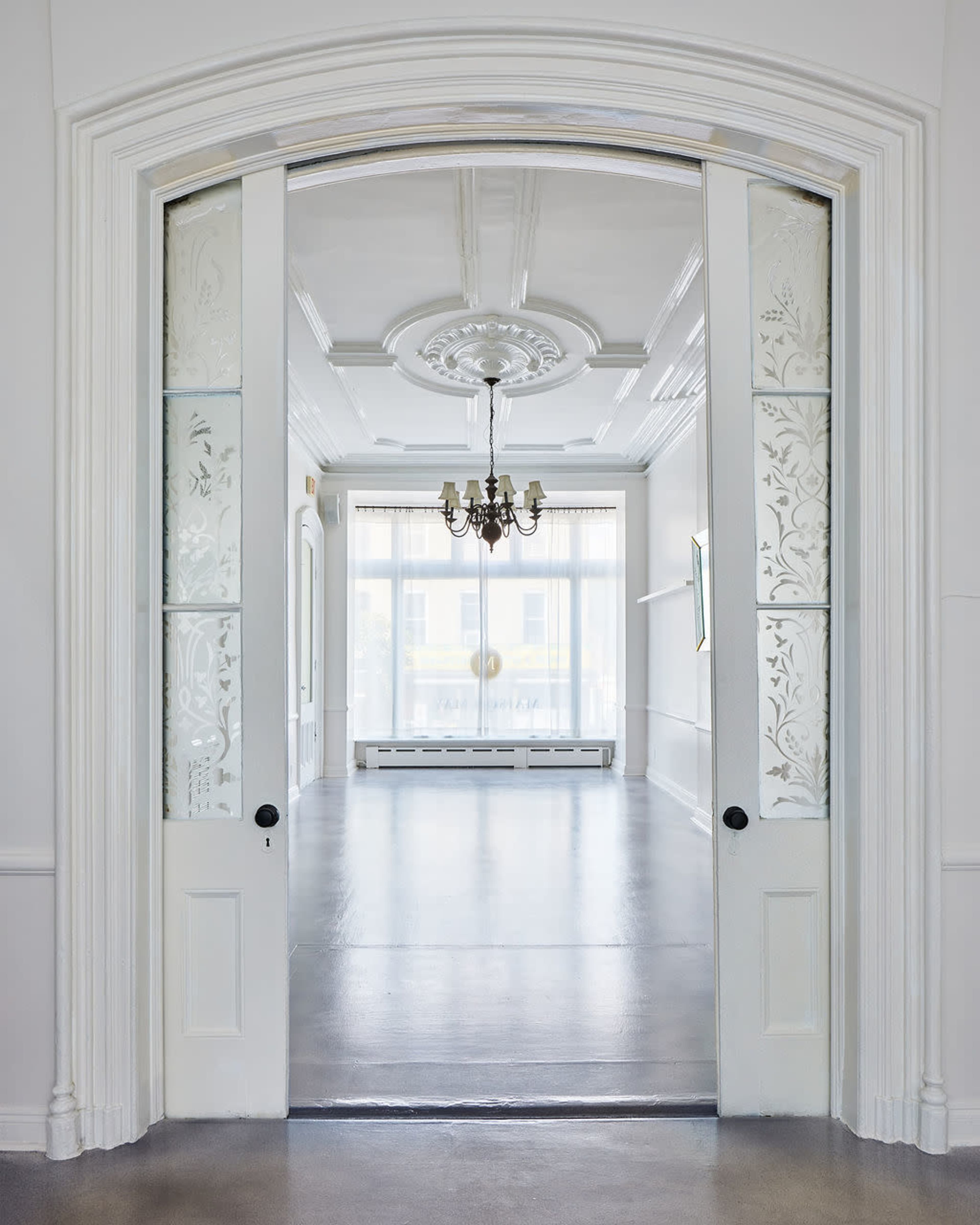 The image shows a spacious, well-lit room viewed through open double doors, with decorative glass panels and a chandelier hanging from the ceiling.
