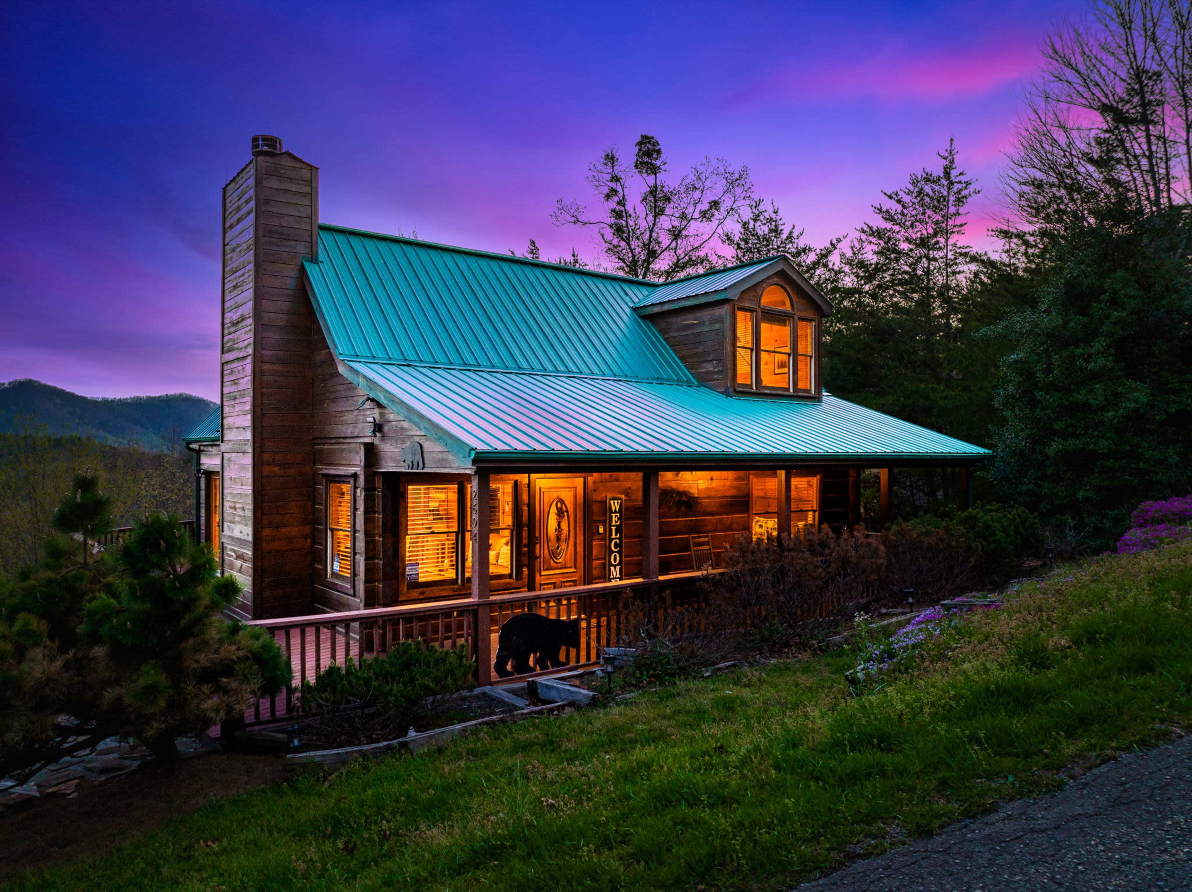 A wooden cabin with a green metal roof, illuminated by warm lights against a purple and blue twilight sky, surrounded by trees and landscaped greenery.