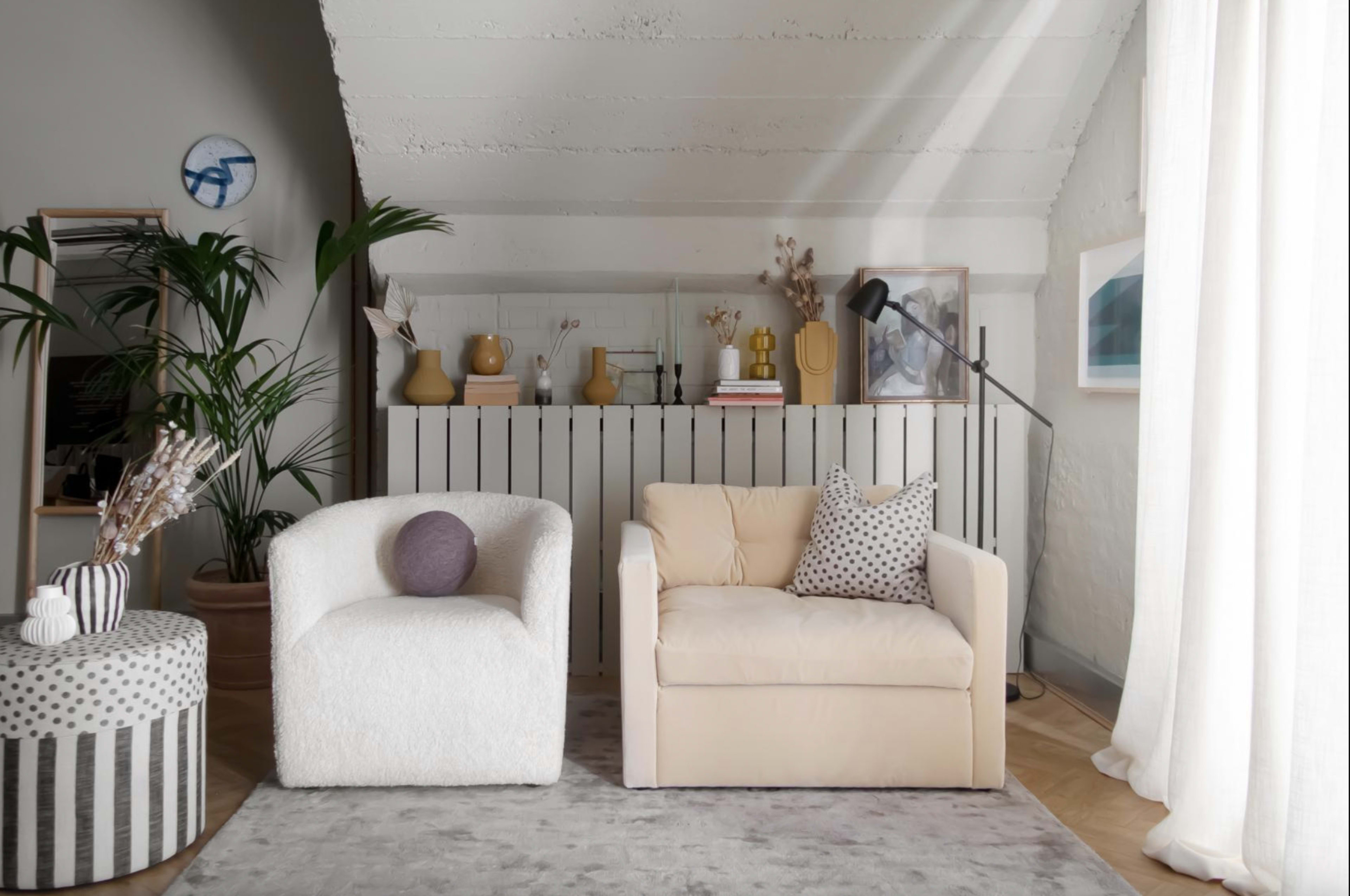 A cozy living room featuring two armchairs, a round striped table, and a light-colored sofa, with plants and decorative objects arranged on a low shelf.
