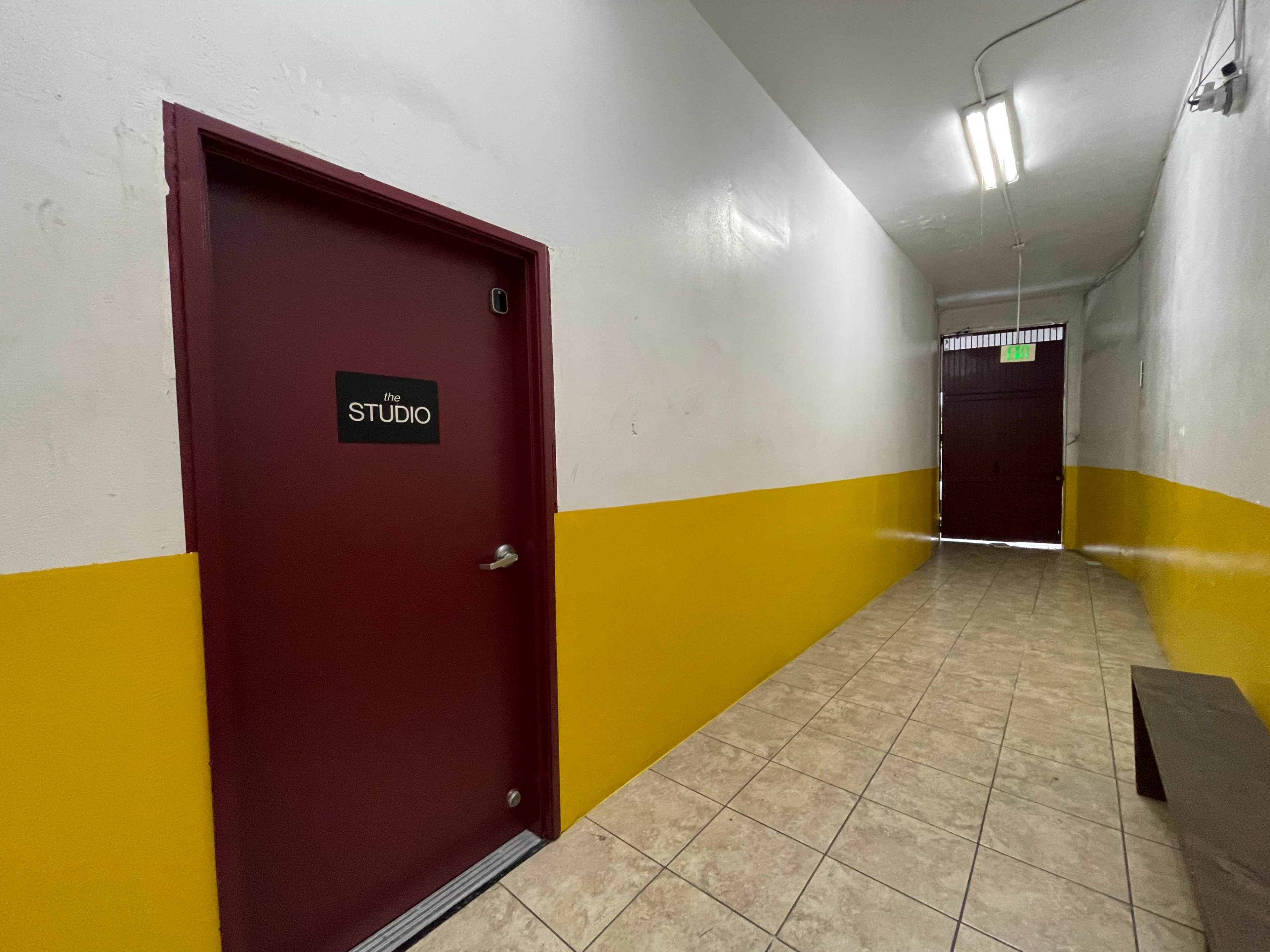 The image shows a narrow hallway with a maroon door labeled "THE STUDIO," flanked by a yellow-painted wall and tiled flooring.