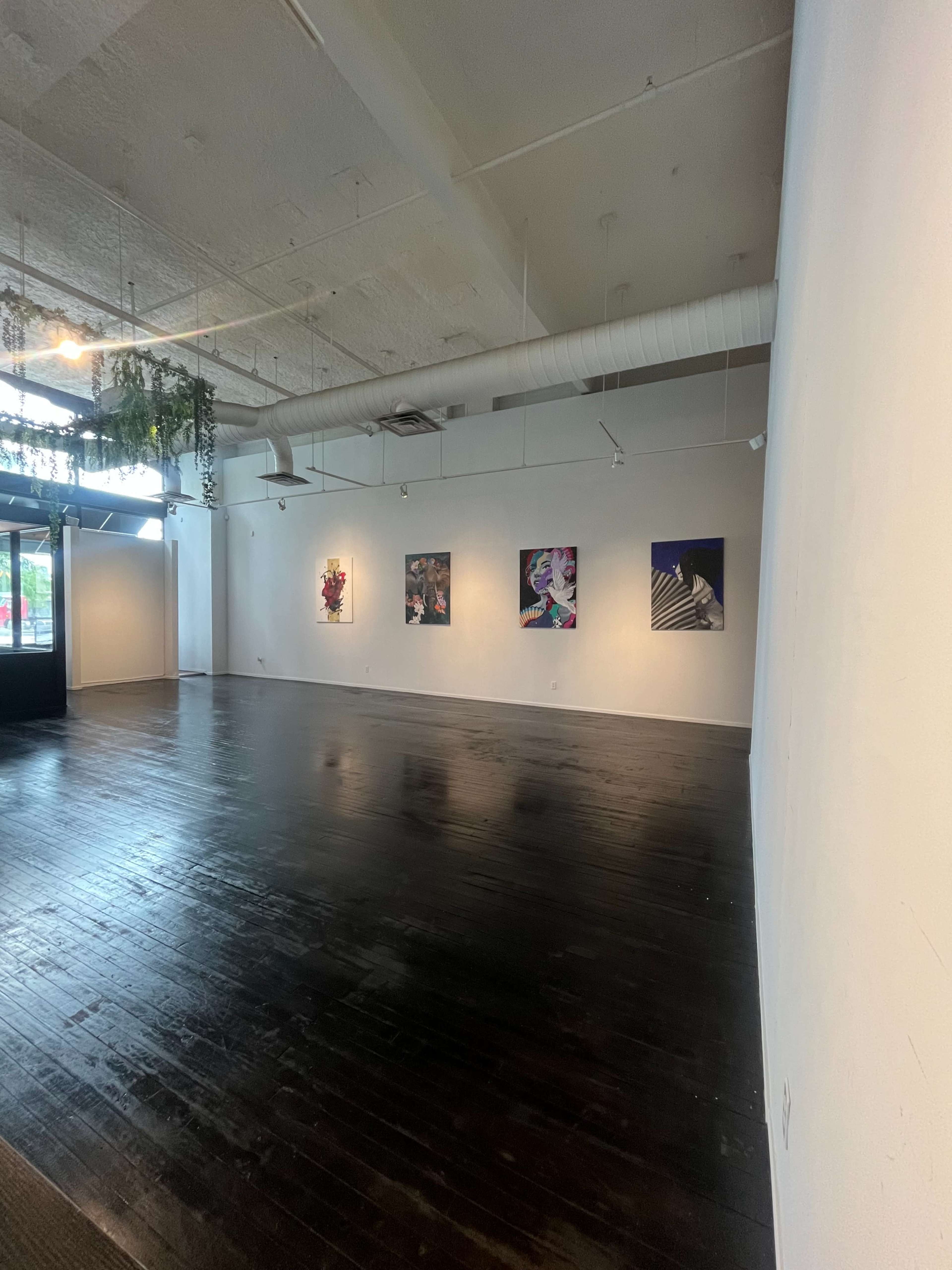 The image shows an empty art gallery space with four paintings displayed on a white wall and dark wooden flooring.
