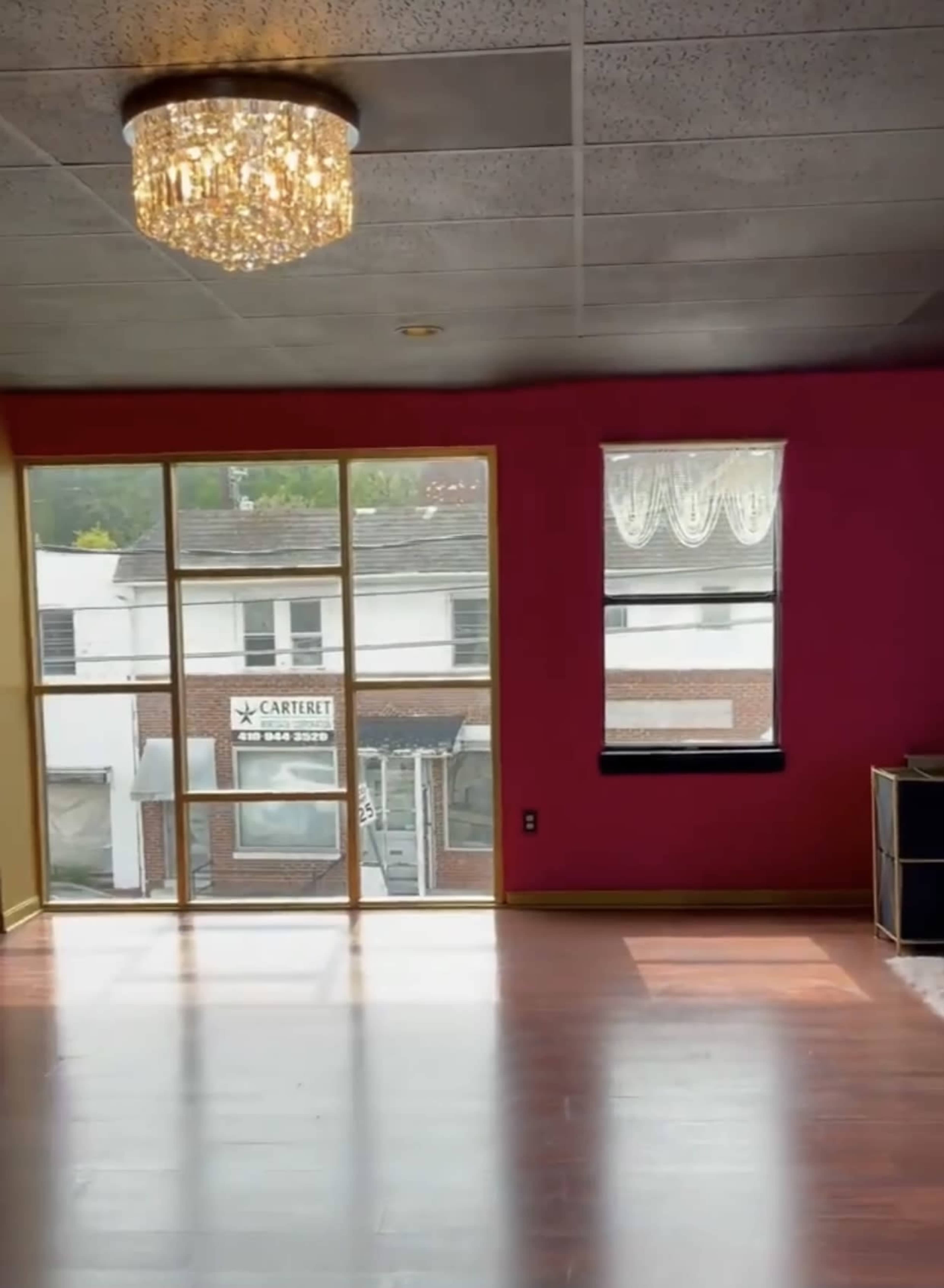 An empty room with polished wooden floors, a pink accent wall, a ceiling chandelier, and large windows facing an exterior street view.