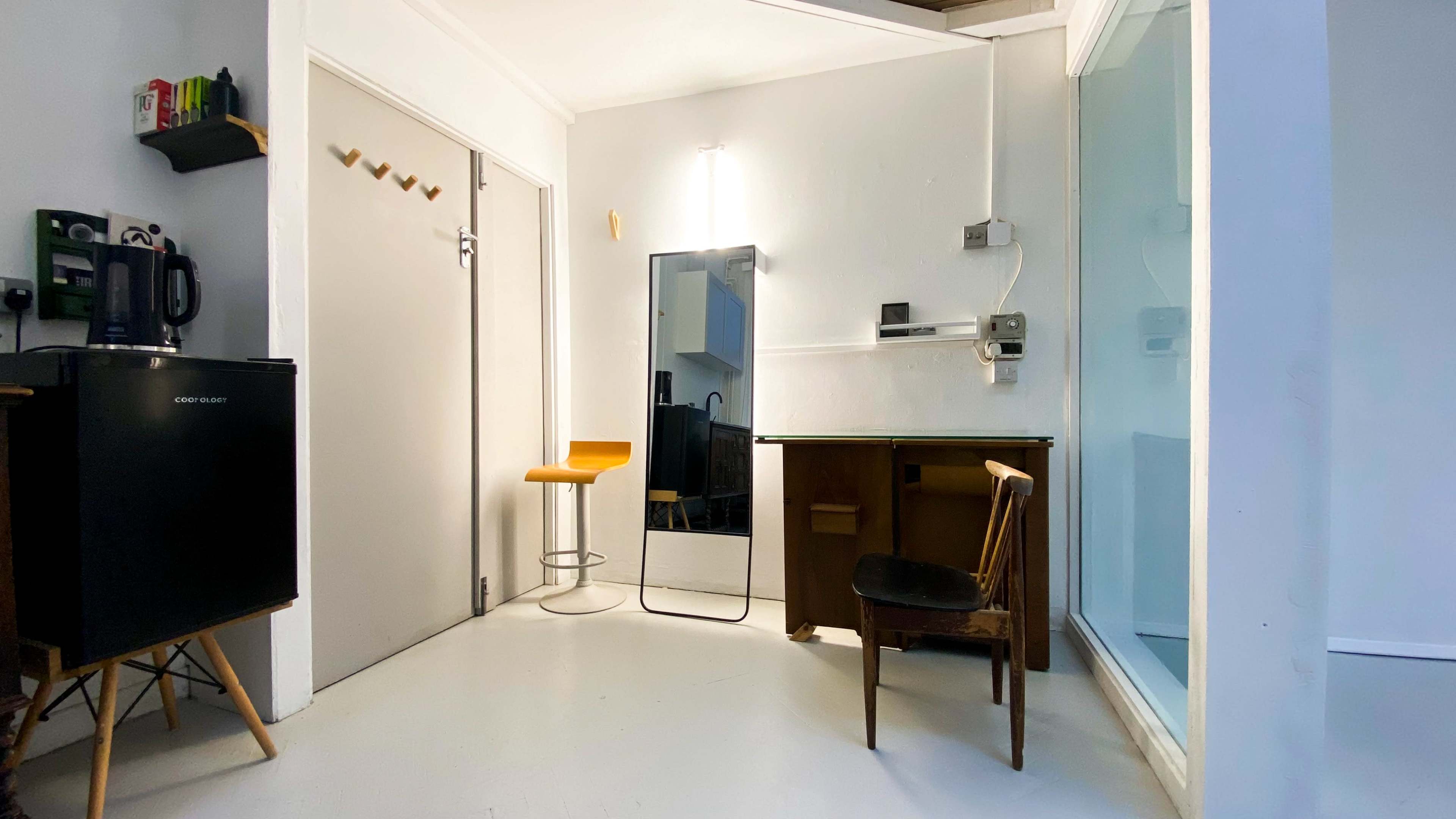 A small room featuring a minimalist workspace with a mirror, a wooden chair, a bar stool, and a kitchenette area with a black refrigerator.