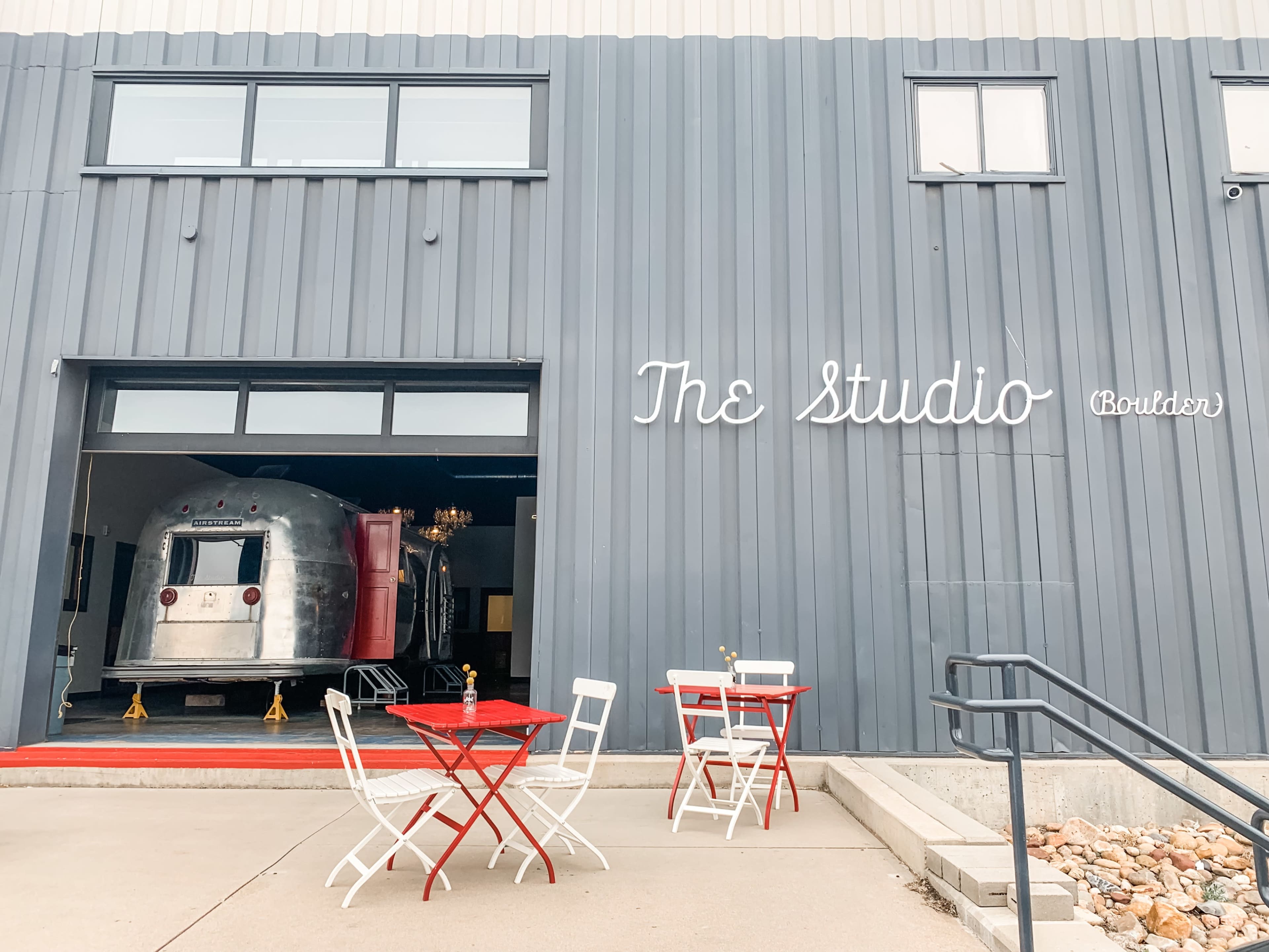 A metal trailer is parked inside a large, open garage with modern signage reading "The Studio" and small red bistro tables in front.