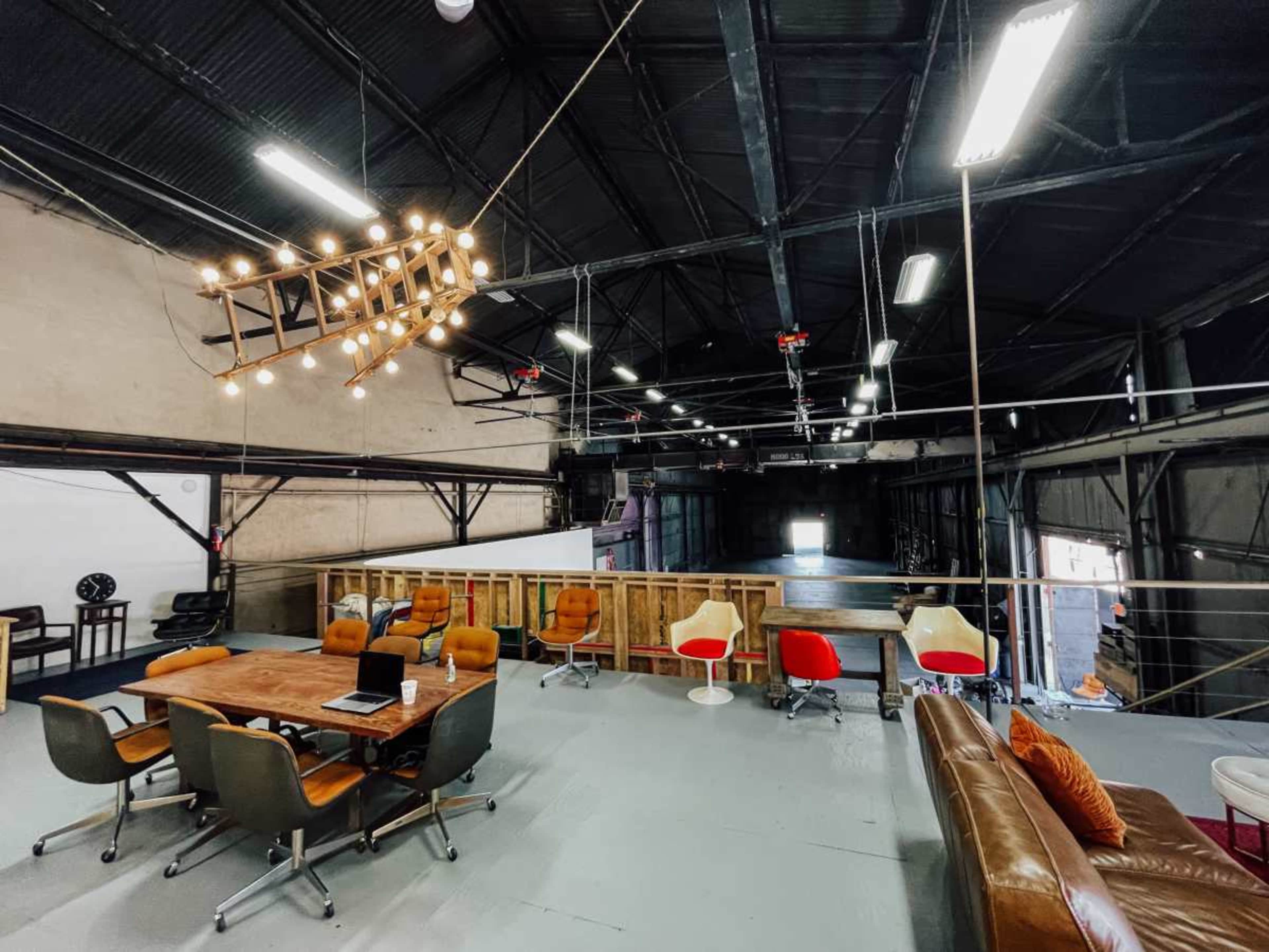 The image shows a spacious, industrial-style loft with a wooden table surrounded by chairs and a sofa, illuminated by a striking chandelier.
