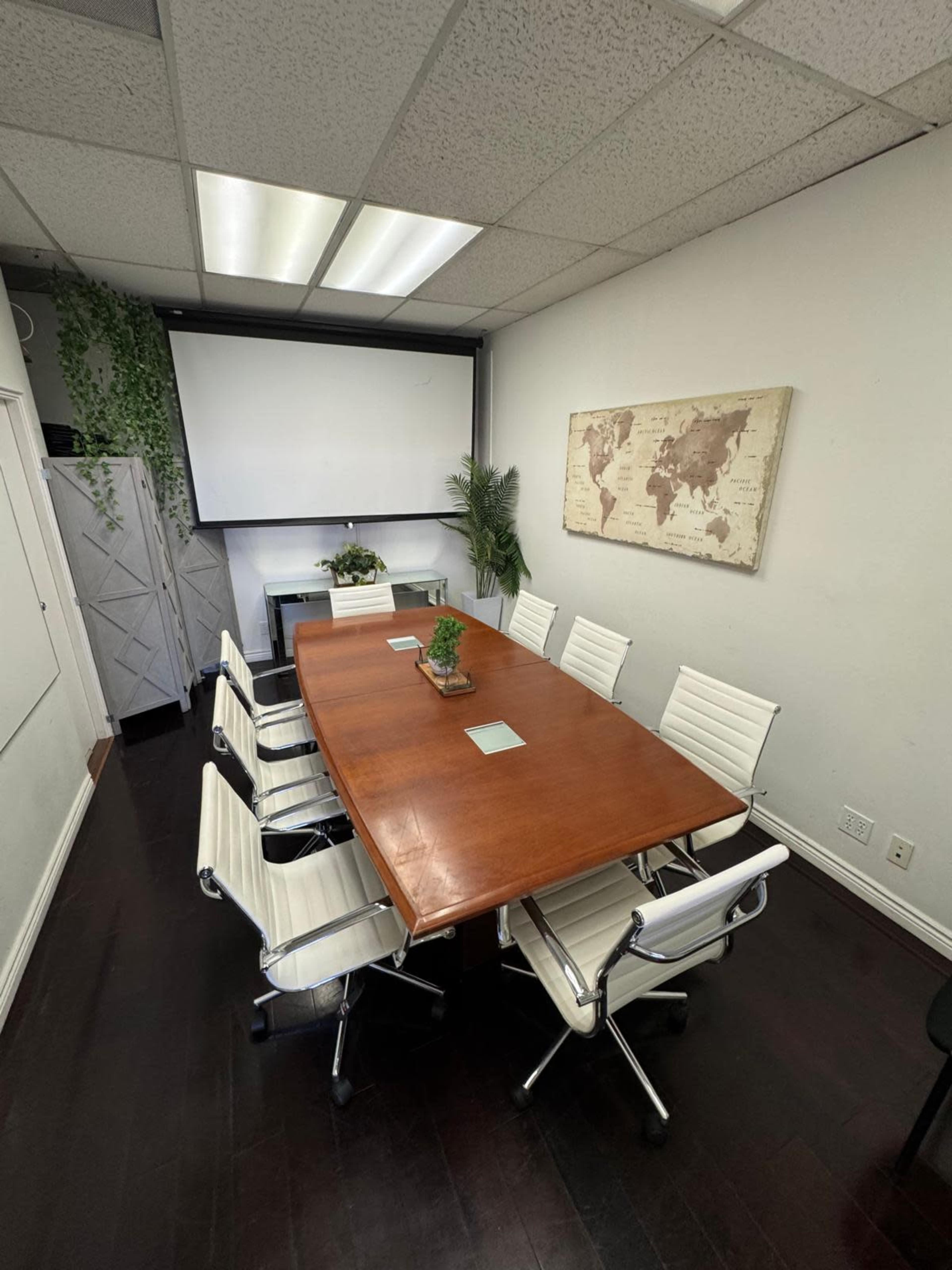 The image shows a conference room with a large wooden table surrounded by eight white chairs, a projector screen on the wall, and a world map hanging decoratively.