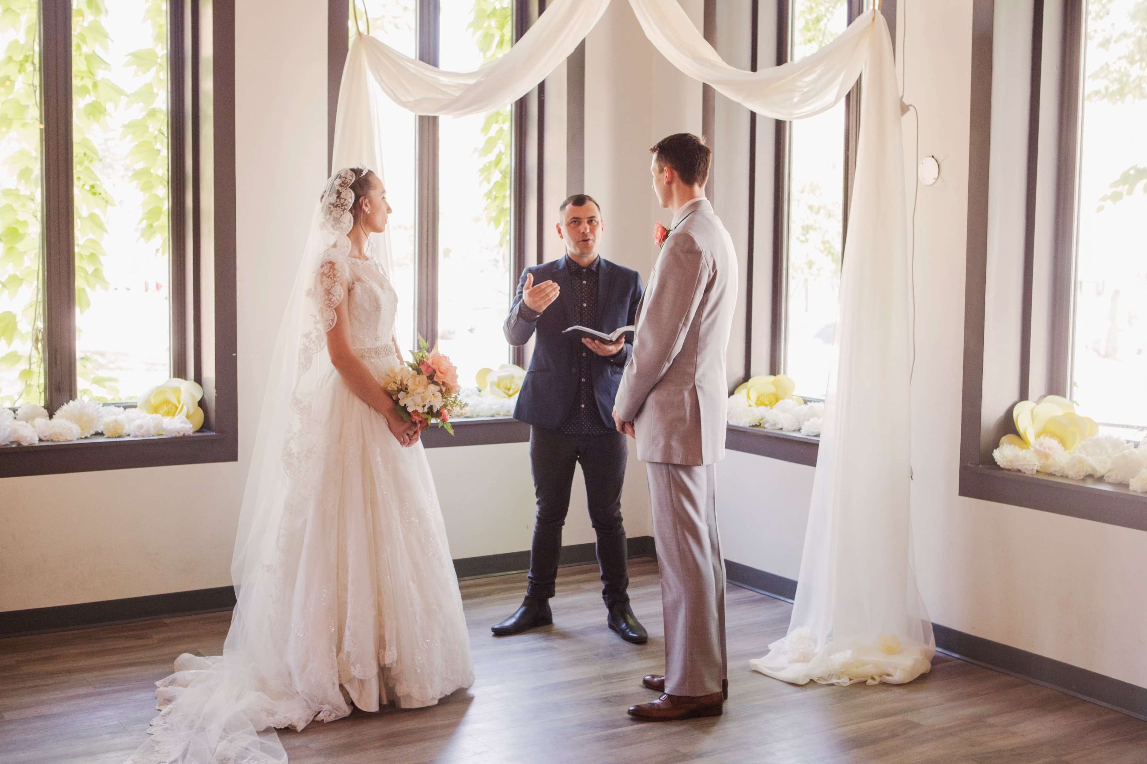 A bride and groom stand beneath a draped fabric arch while a officiant addresses them during a wedding ceremony.