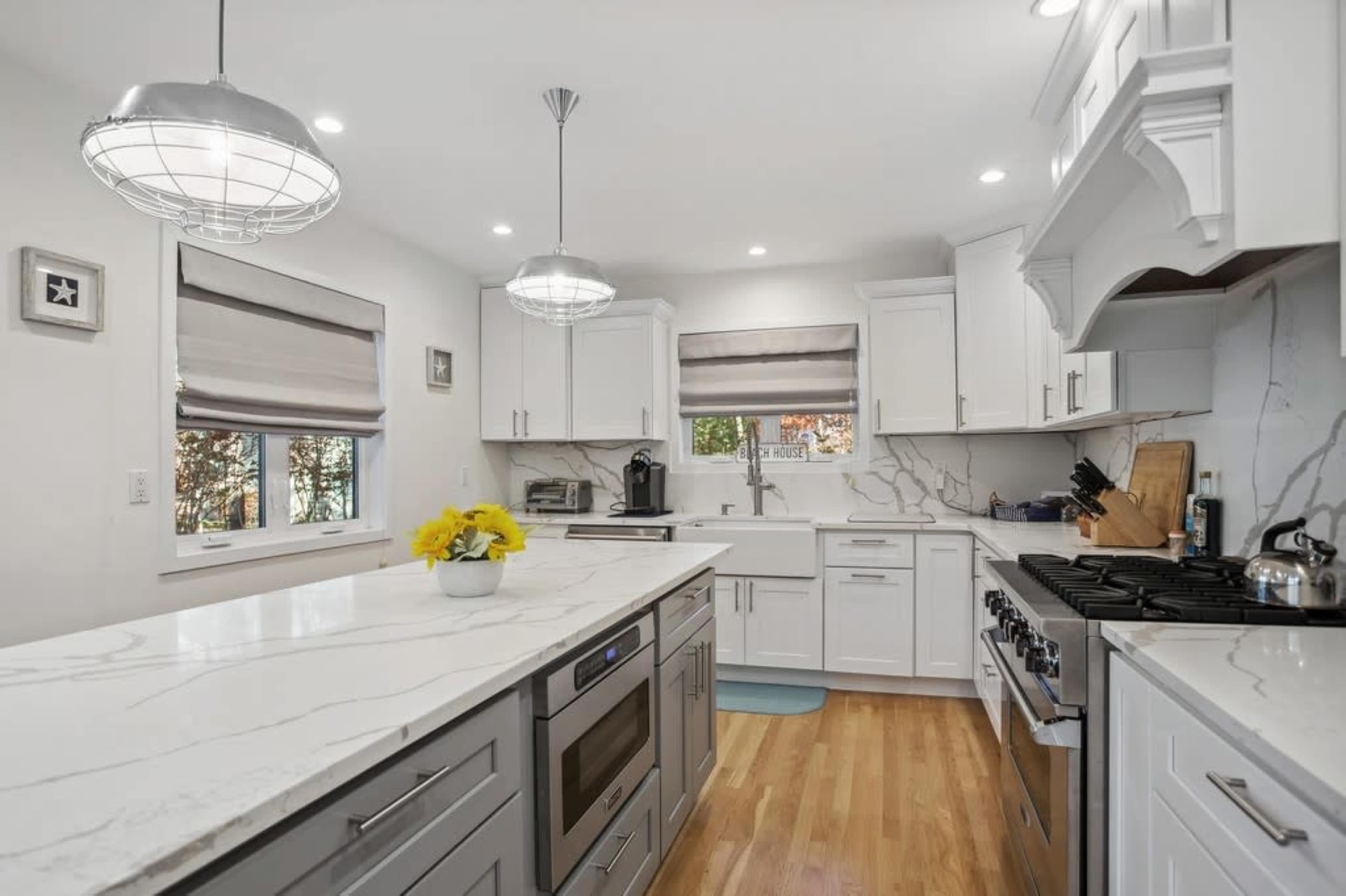 A modern kitchen features white cabinets, a marble countertop, and stainless steel appliances, with pendant lighting and large windows providing natural light.
