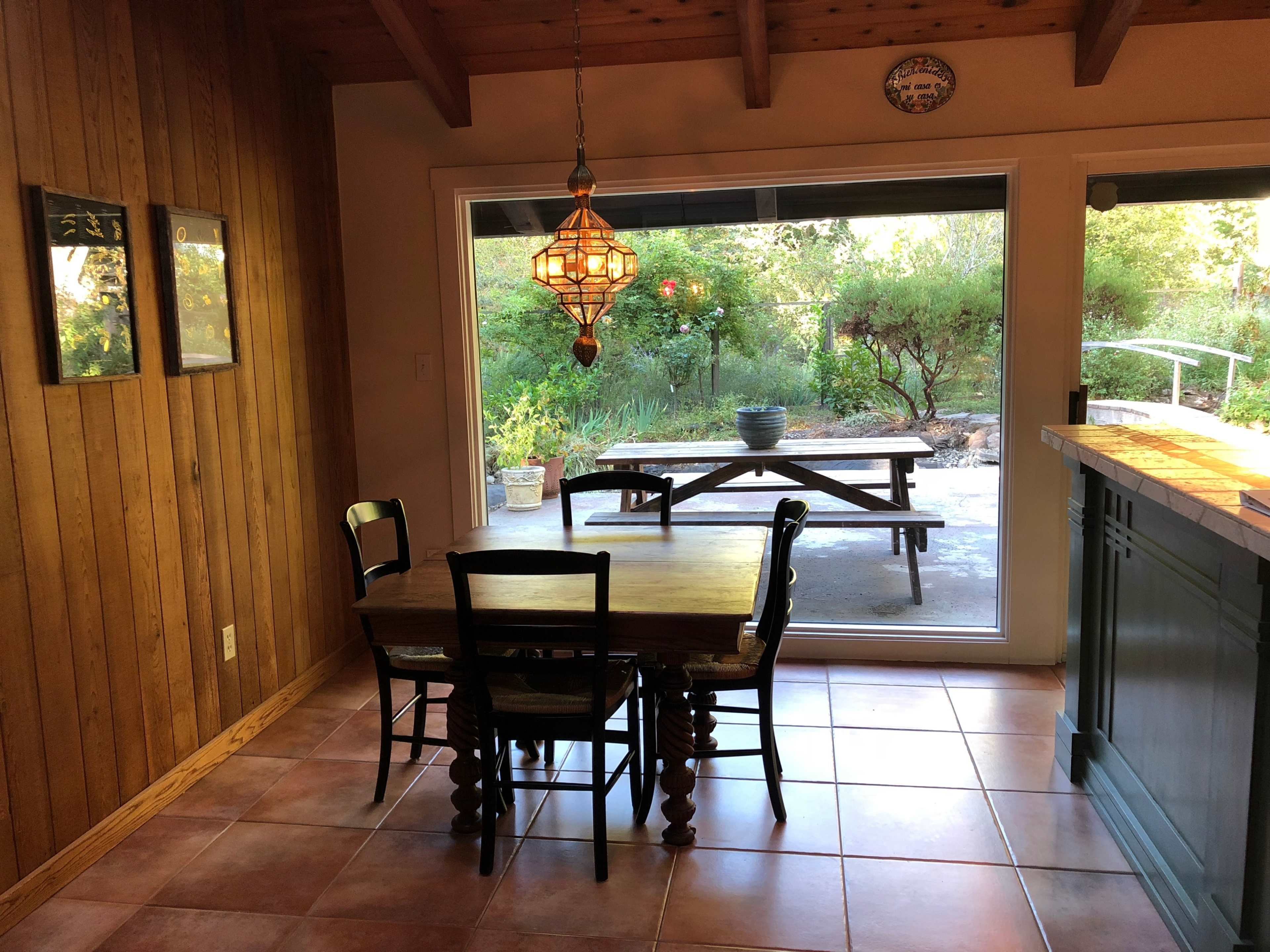 A wooden dining table with four chairs is situated inside a room that opens to a patio with a view of greenery and outdoor seating.