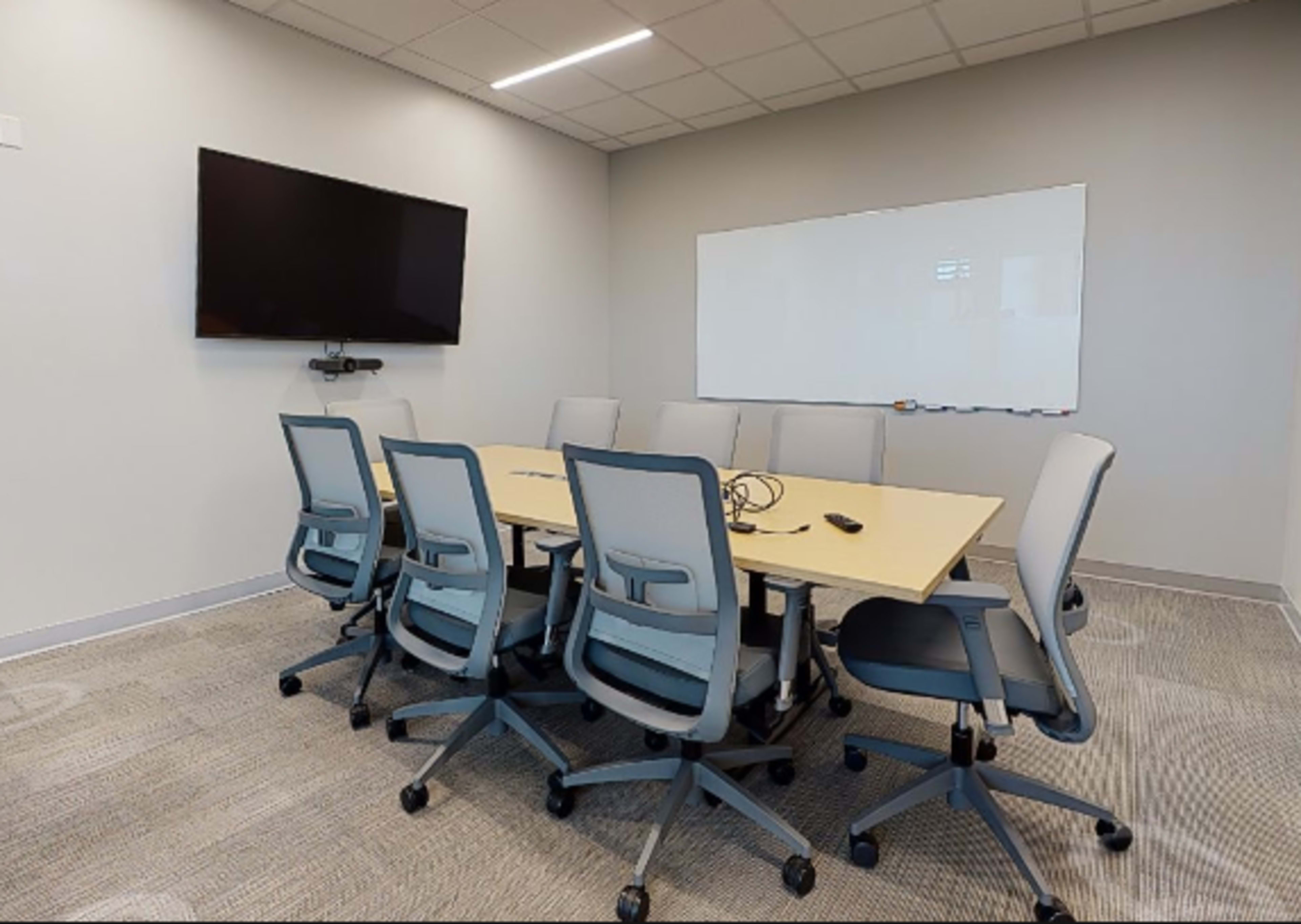 A conference room is equipped with a large table surrounded by eight chairs, a television mounted on the wall, and a whiteboard.