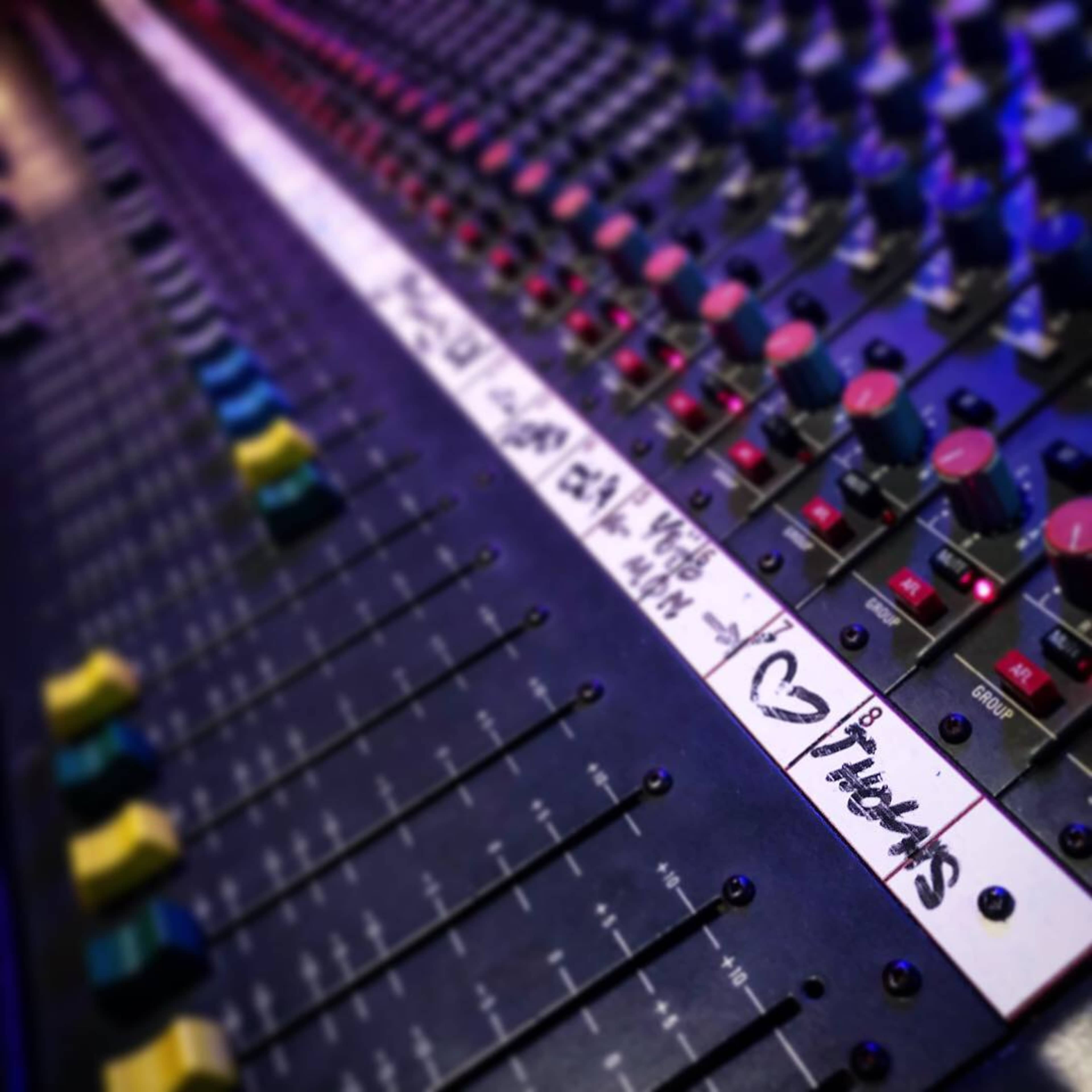 The image shows a close-up of a recording studio mixing console with various colorful knobs and labeled tracks.