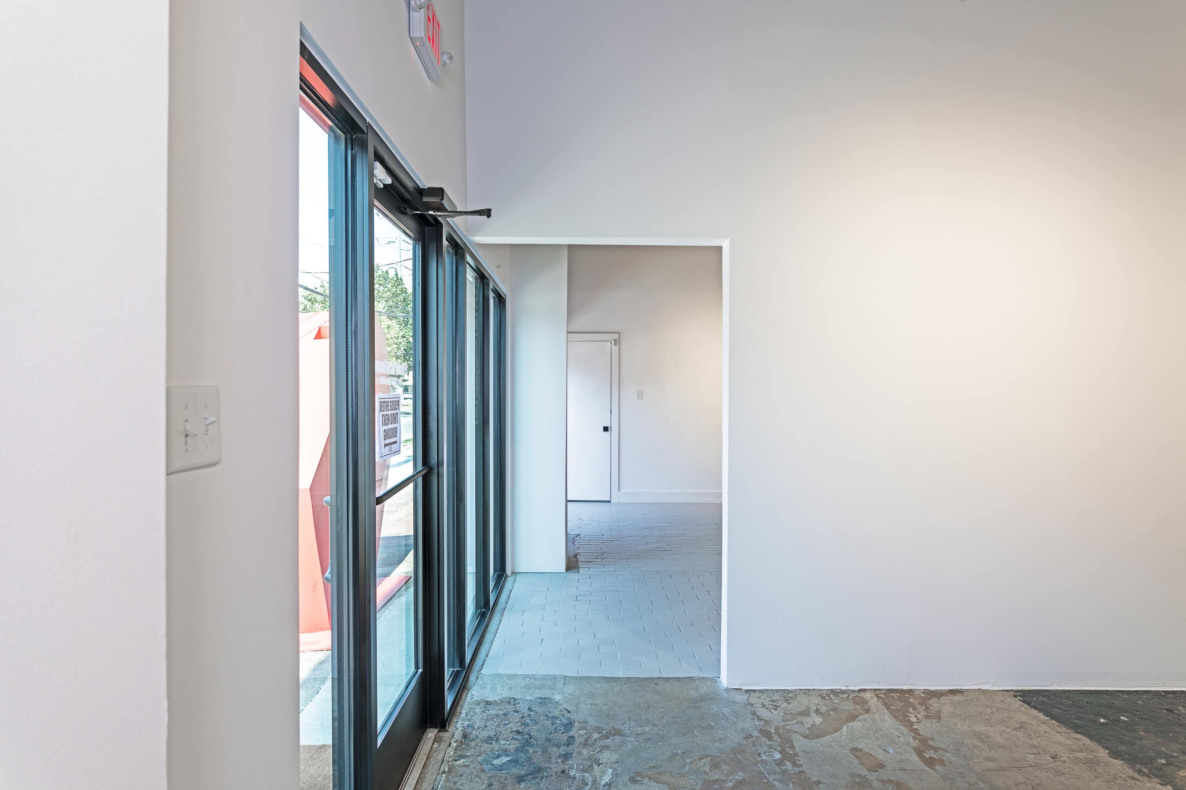 A bright interior space with an open doorway leading to another room, featuring large glass doors on the left and plain walls.