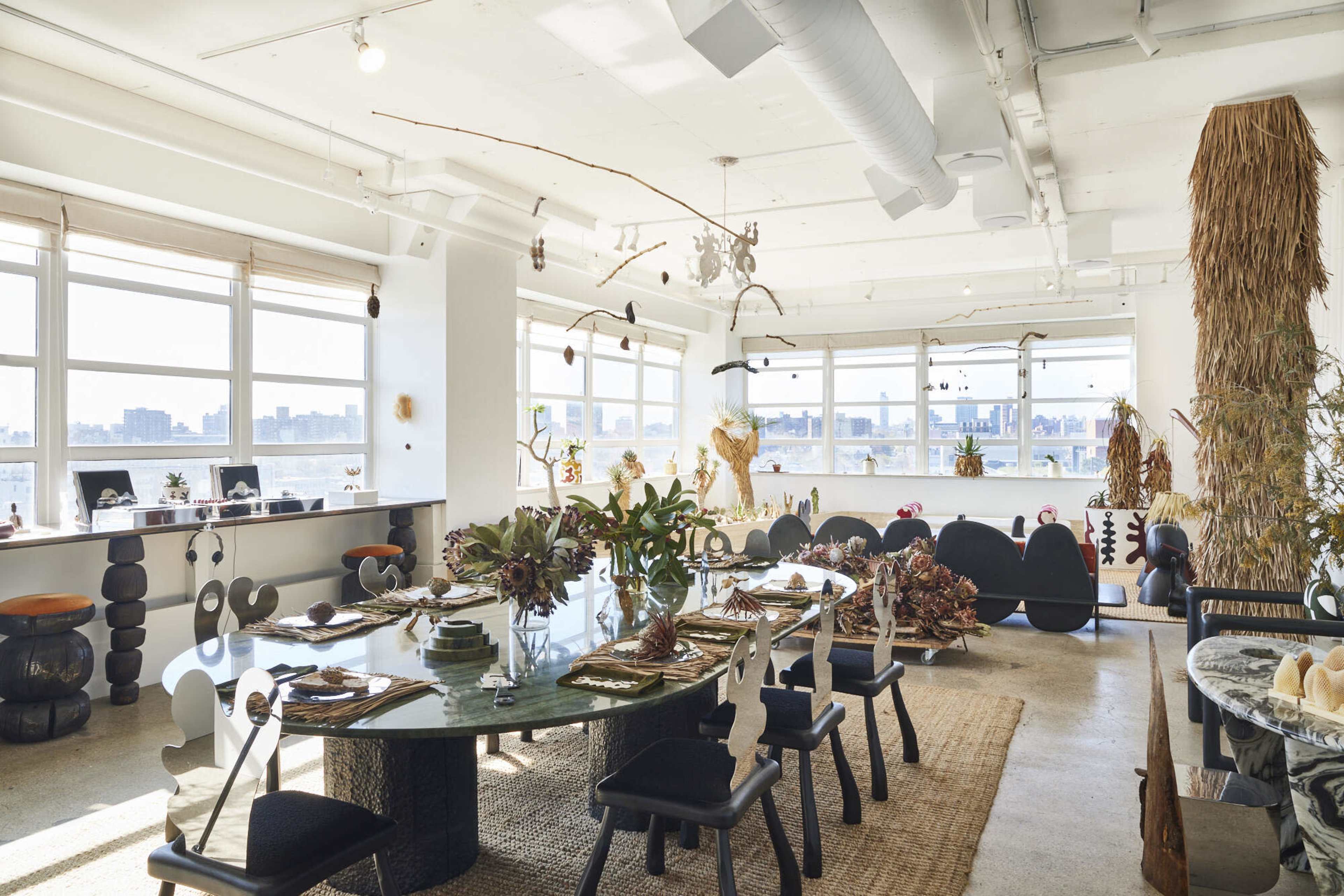 The image shows a spacious, well-lit dining area featuring a large circular table surrounded by various chairs, with artistic decor elements and views of a city skyline visible through large windows.