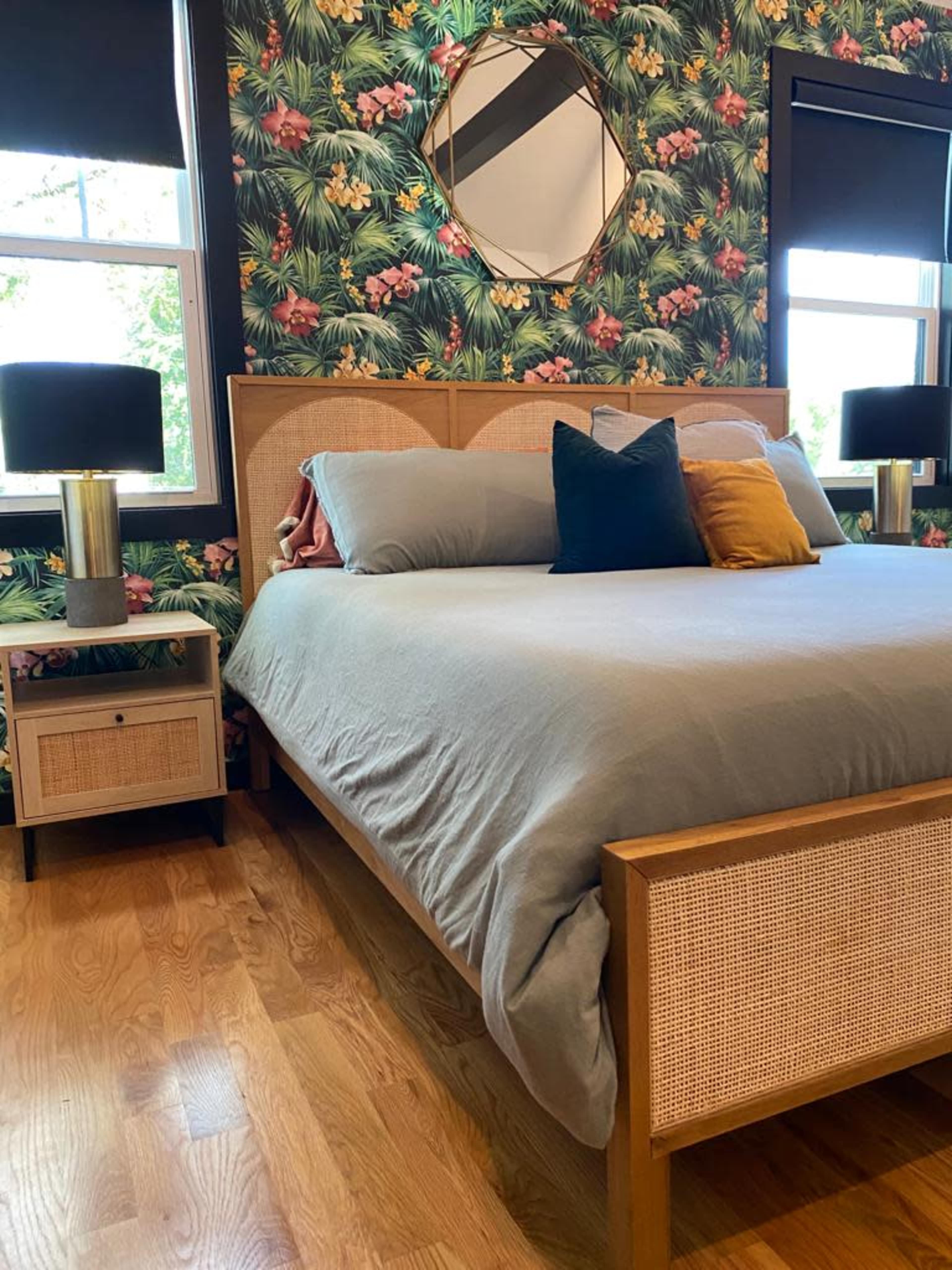 A neatly arranged bedroom featuring a bed with gray bedding, a bedside table, and a floral wallpaper design with tropical leaves and flowers.