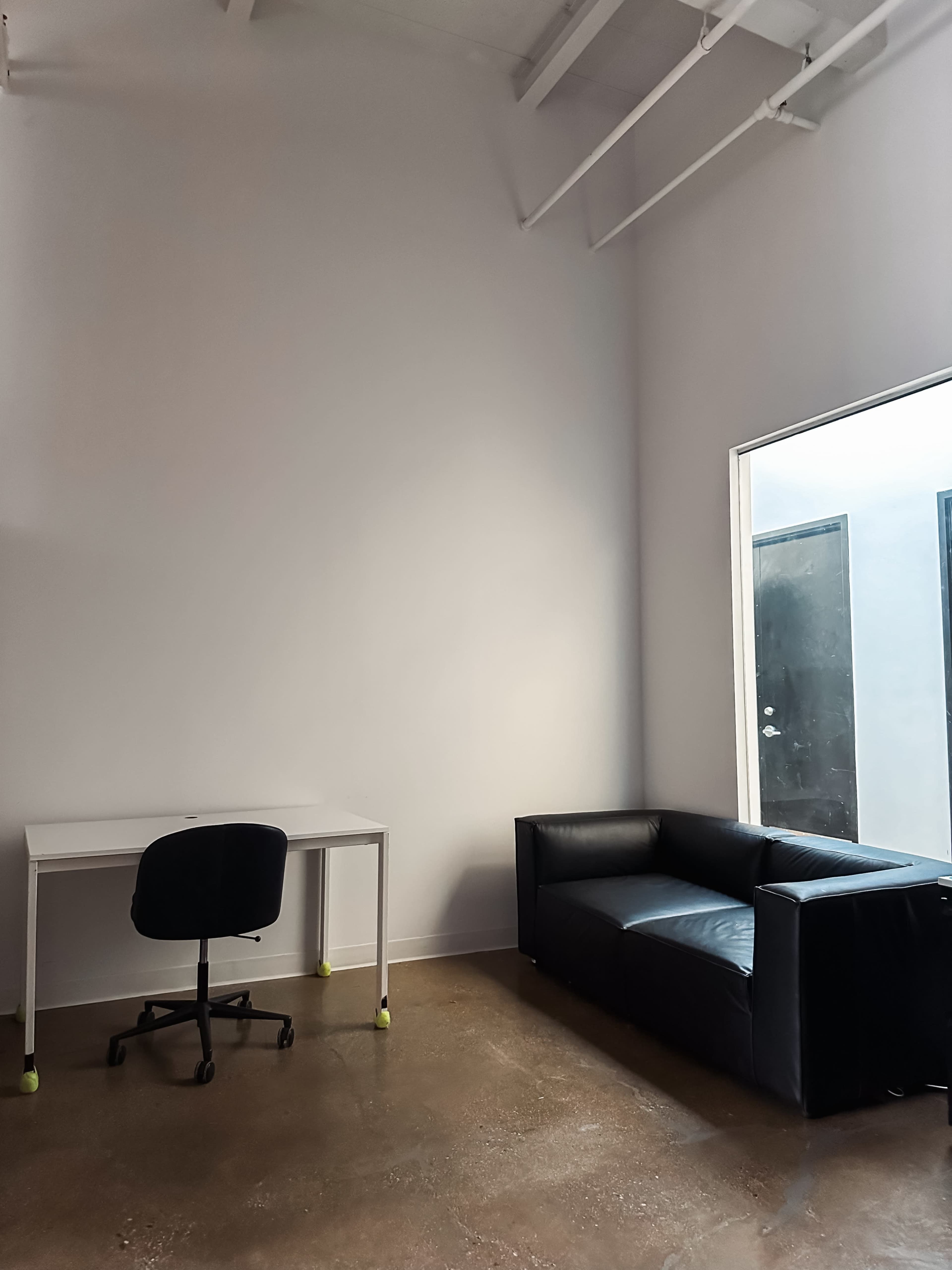 A minimalistic room with a white wall, a small desk, an office chair, and a black couch, with a large window on one side.