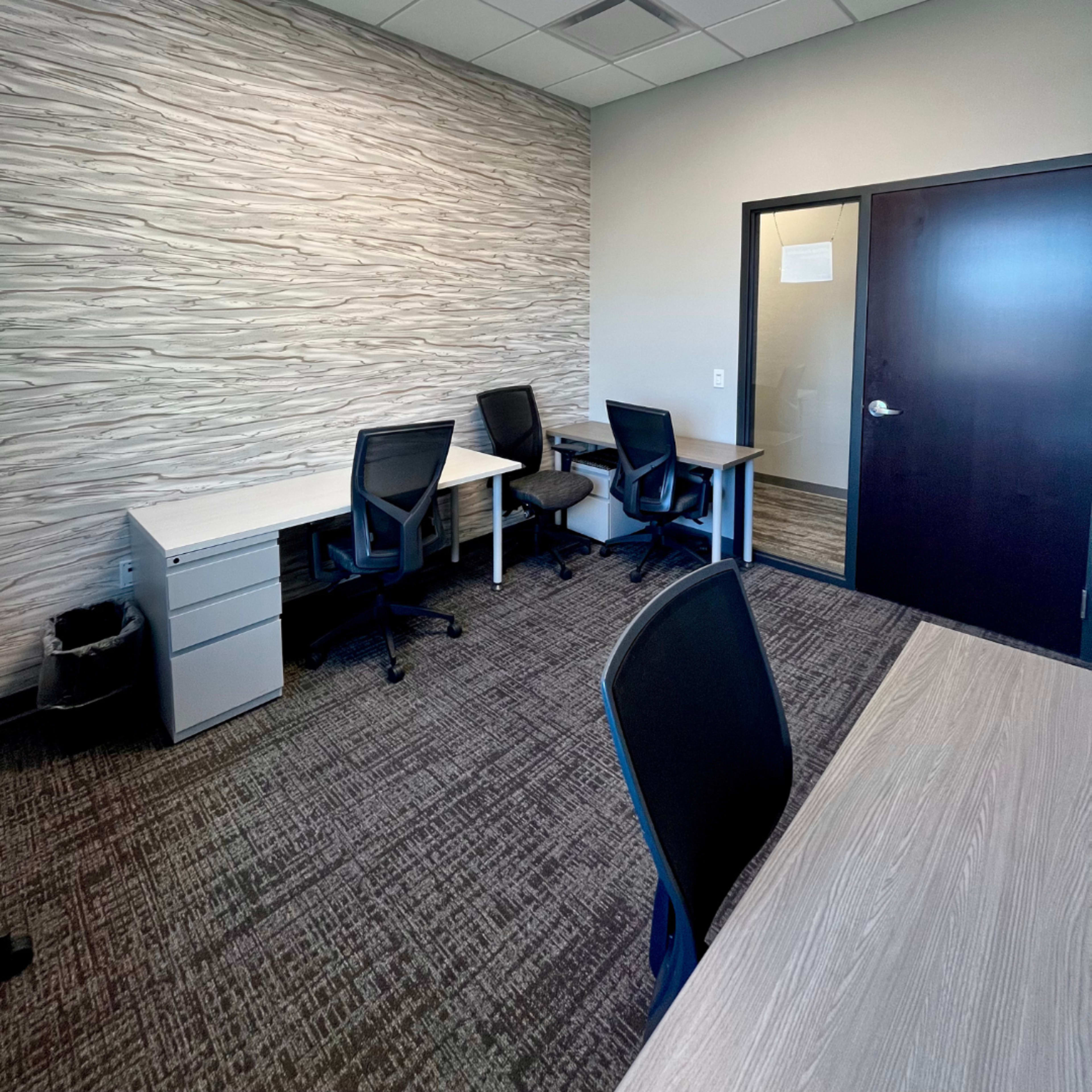 The image shows a modern office space with two desks and chairs against a textured wall, and a door leading to another room.