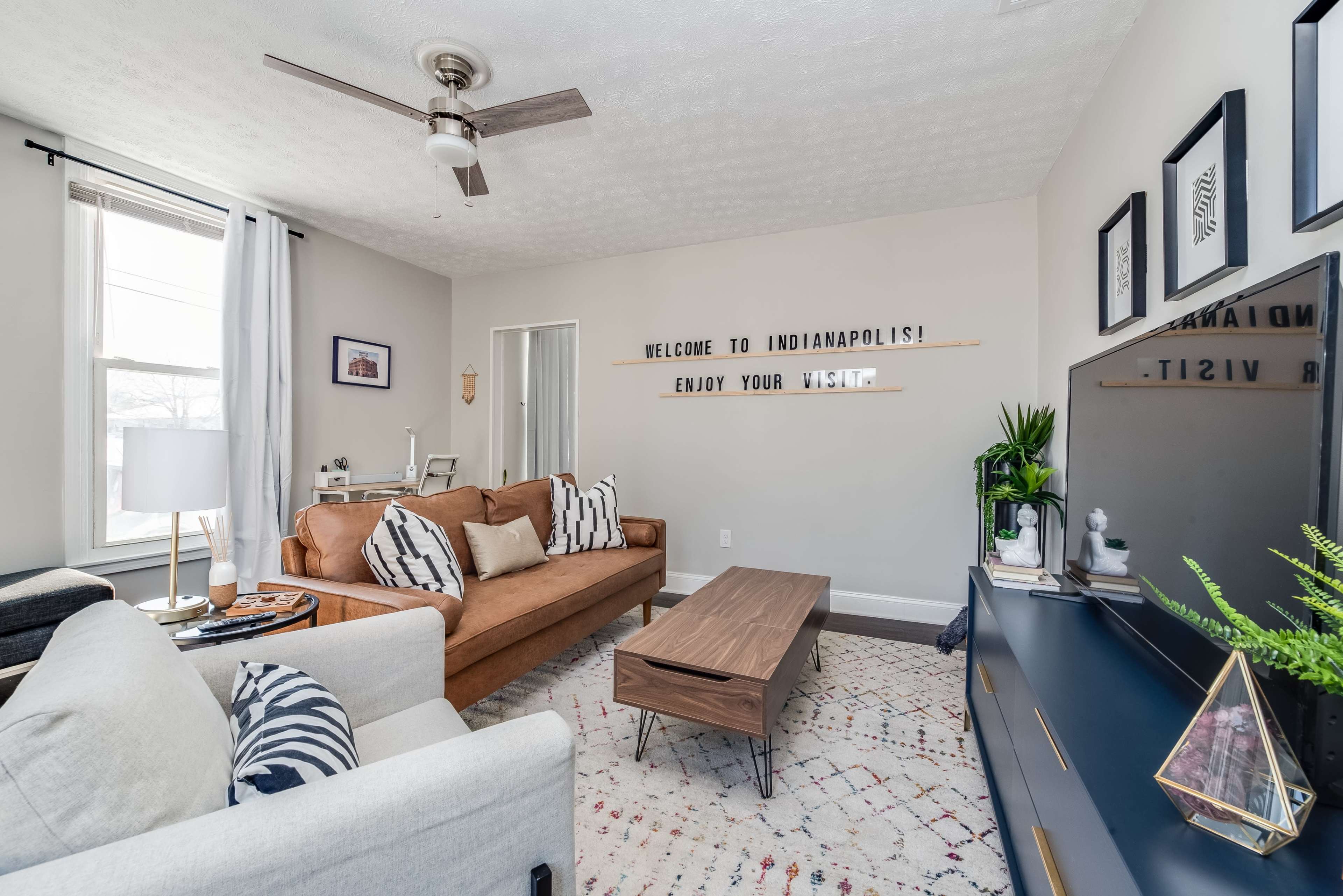 The living room features a brown sofa, a coffee table, and a television against a light-colored wall, with decorative signs and plants enhancing the space.