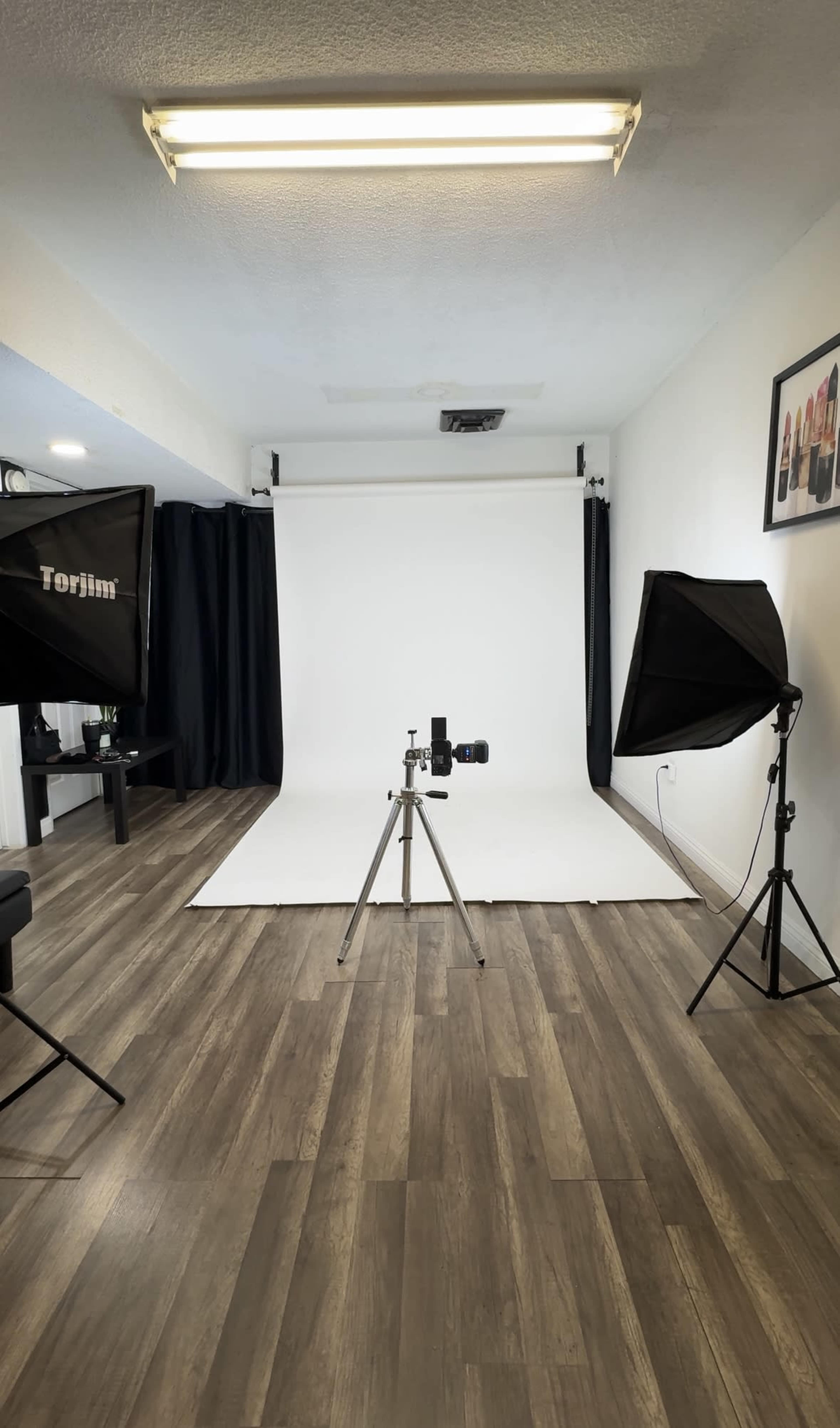 The image shows a photography studio with a white backdrop, a camera on a tripod, and two softbox lights positioned on either side.