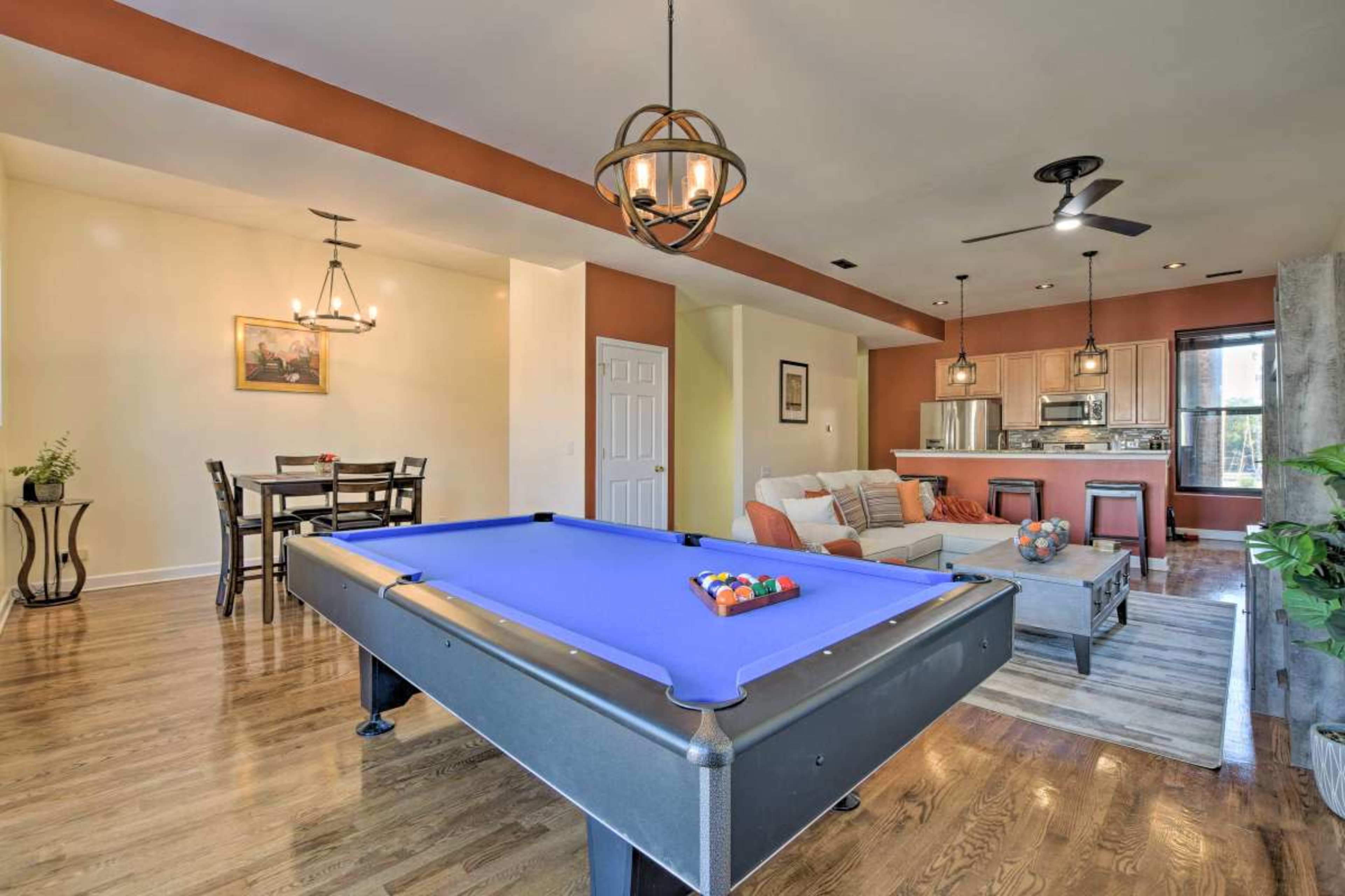 A modern living space with a pool table in the foreground, a dining area with a table and chairs, and a kitchen visible in the background.