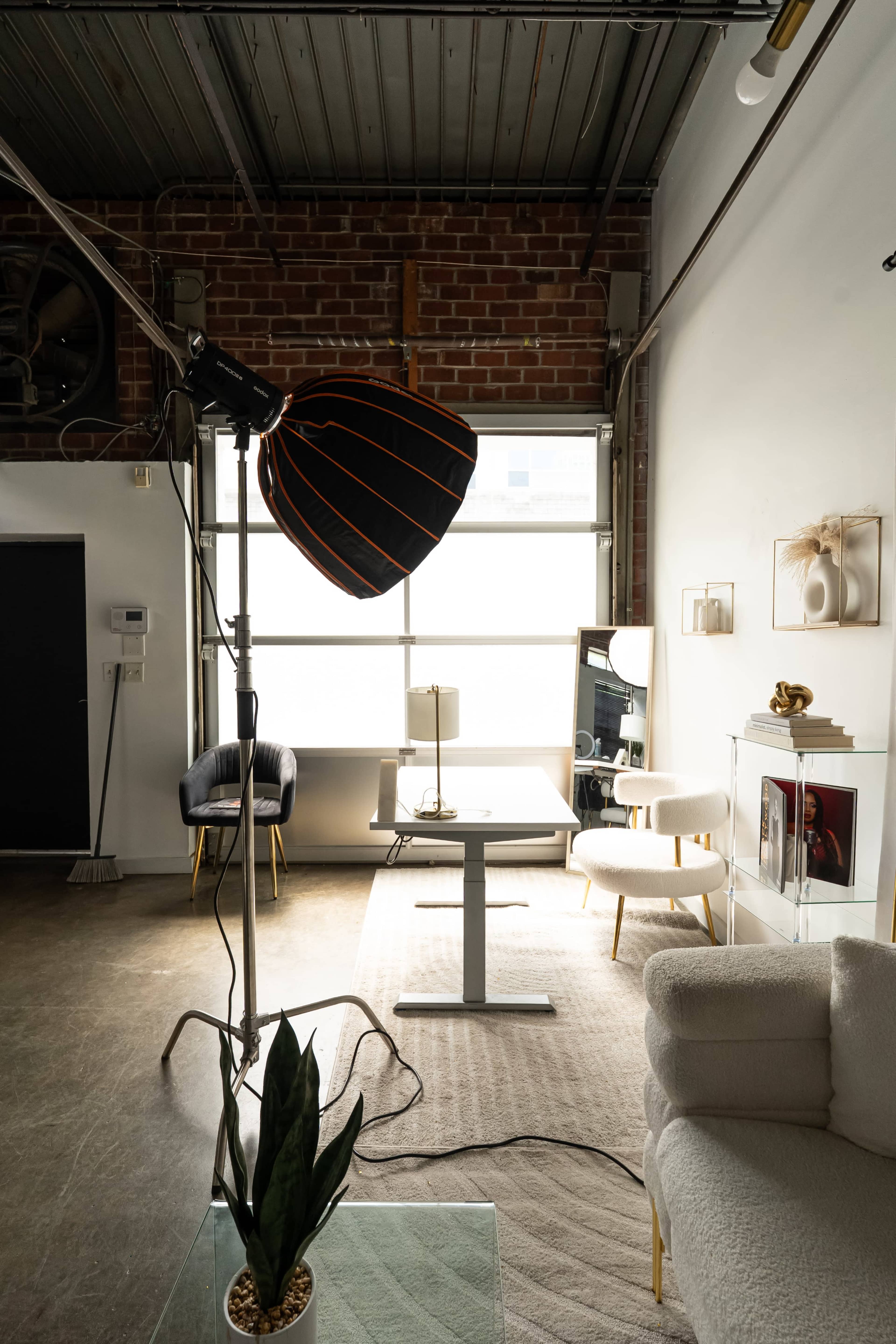 The image shows a modern workspace with a cozy seating area, featuring a large light fixture, a desk, and decorative elements against a brick wall.